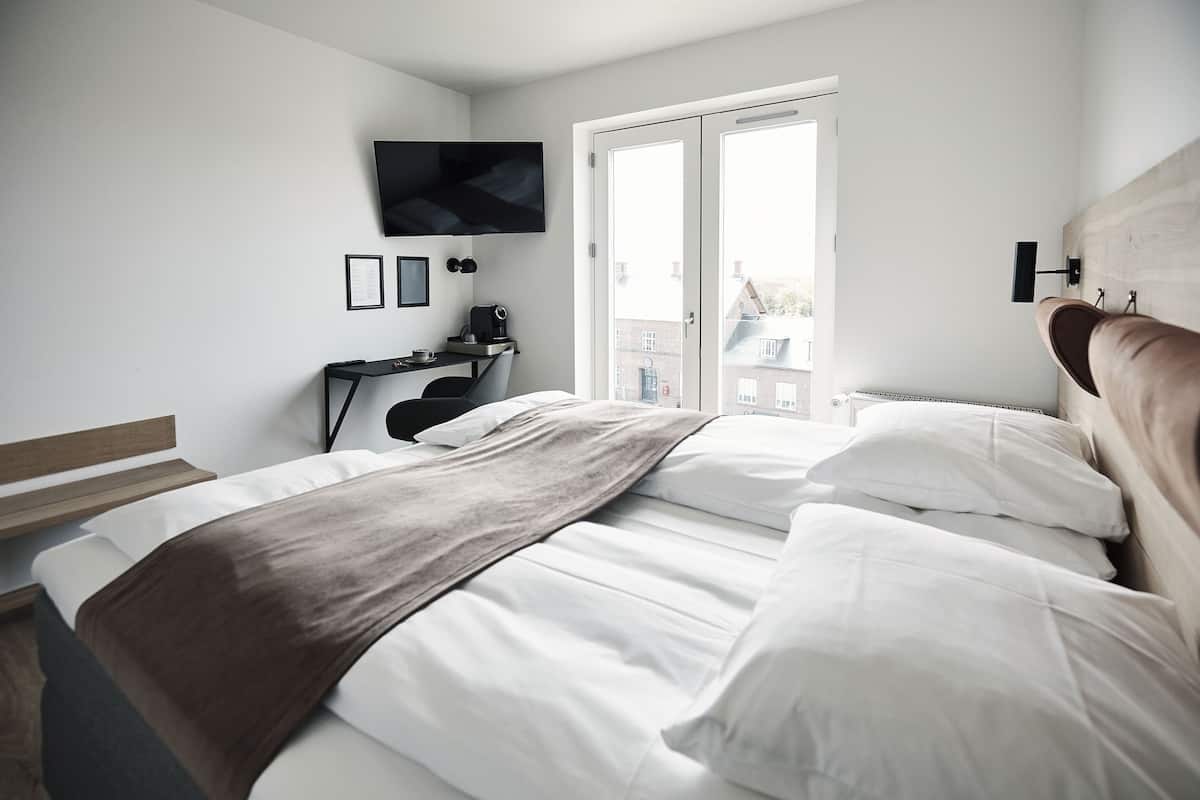 Una habitación de hotel moderna con una cama, un escritorio, un televisor montado en la pared y vistas de la ciudad a través de la ventana.