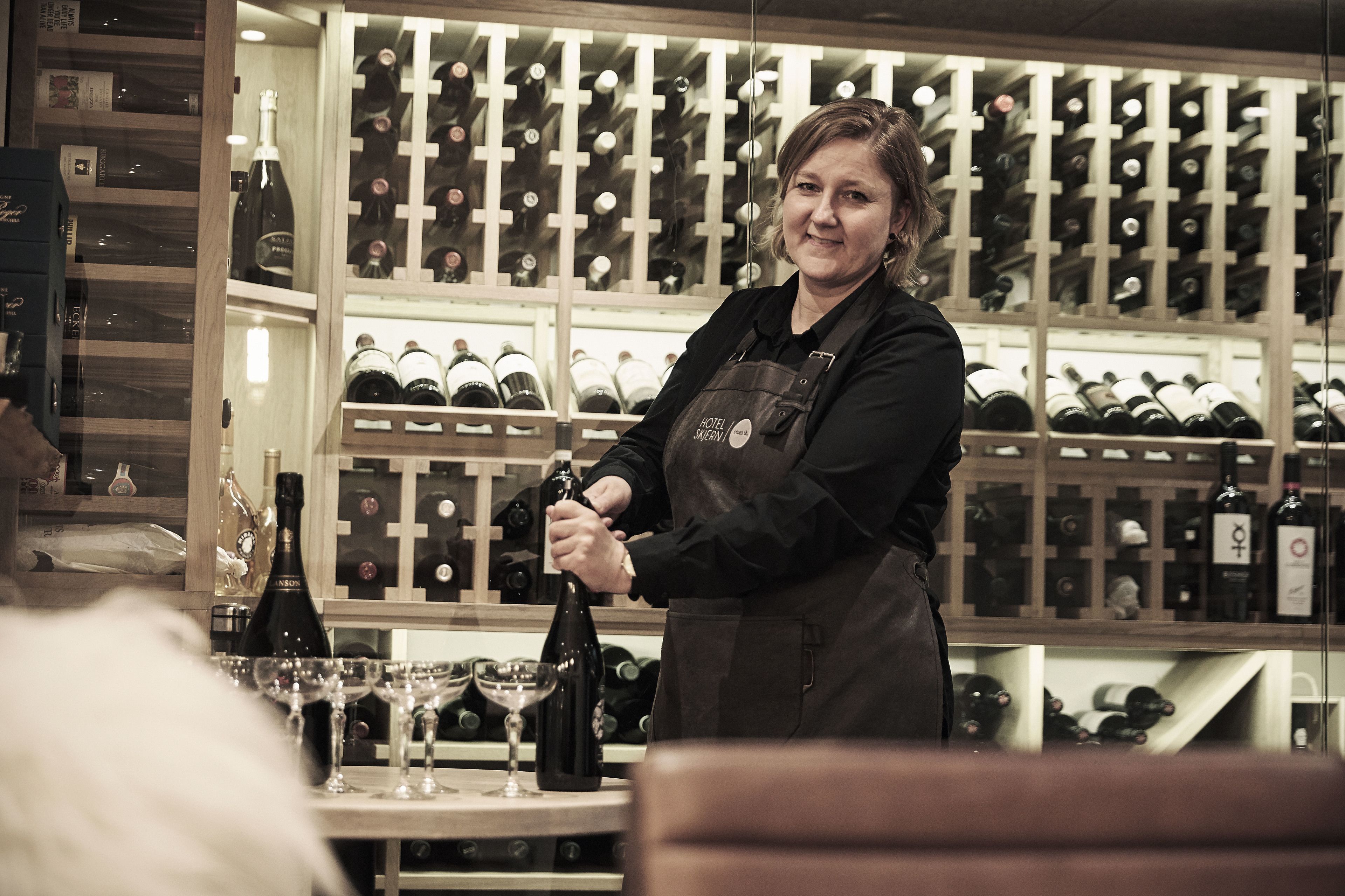 A person in an apron at a bar with a wine rack in the background.