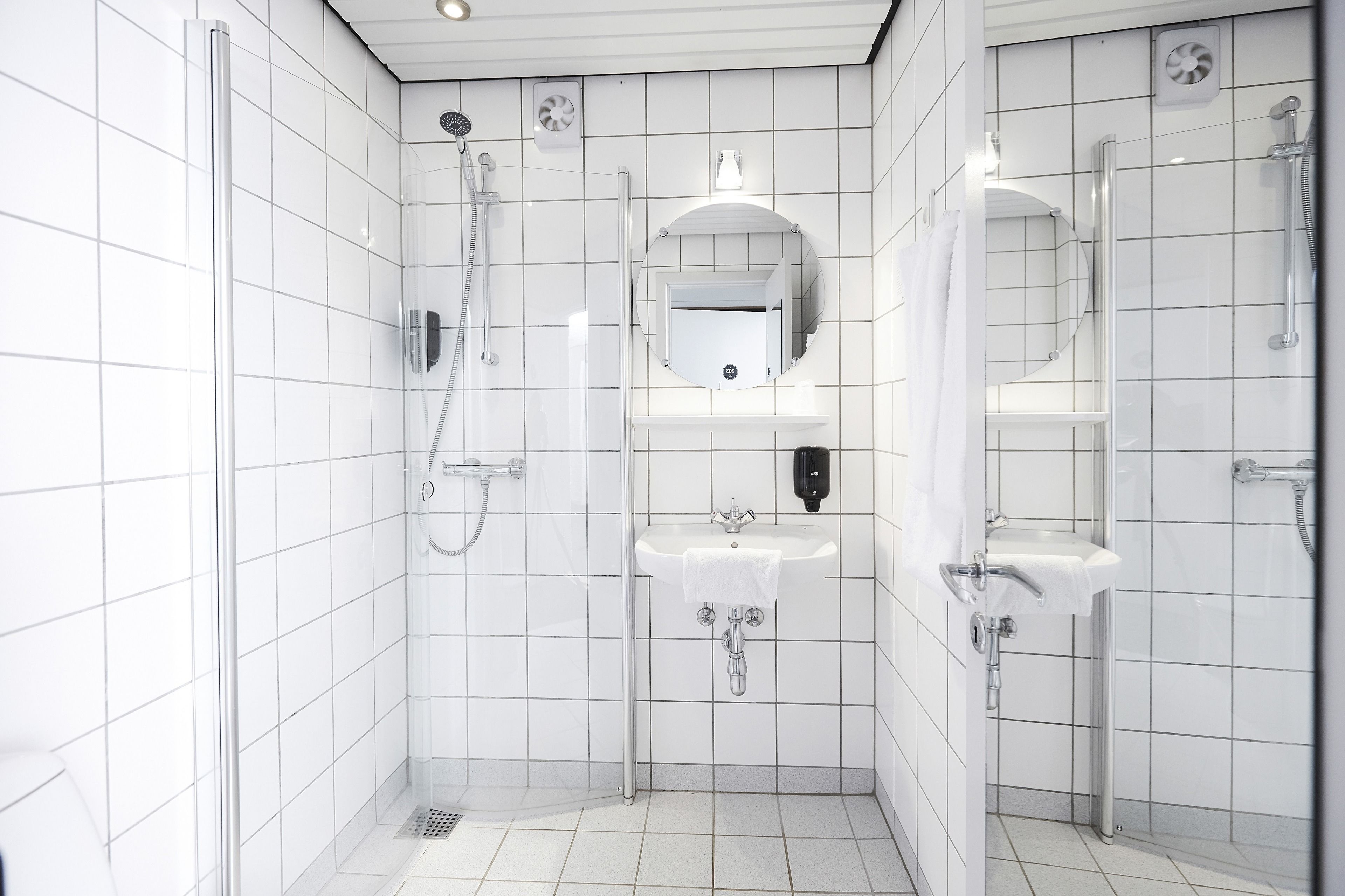 A modern bathroom with white tiled walls, a glass shower enclosure, two sinks, and a mirror.