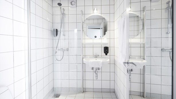 A modern bathroom with white tiled walls, a glass shower enclosure, two sinks, and a mirror. - Hotel Skjern (Skjern)