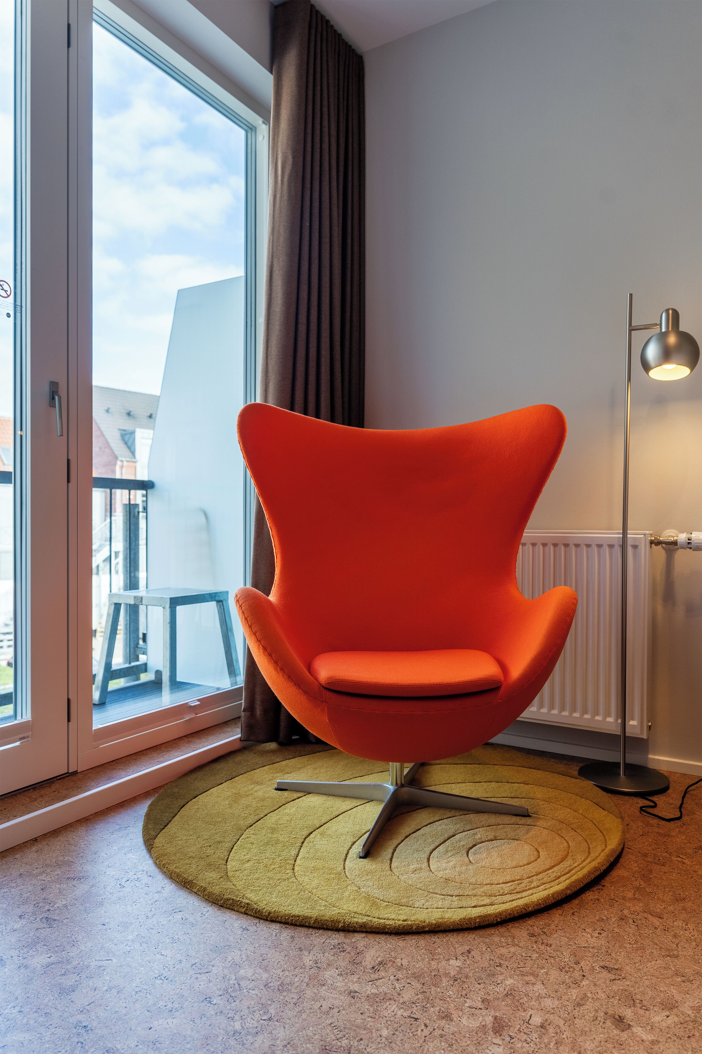 Una habitación moderna con un ventanal grande, un sillón naranja brillante, una lámpara de pie y una mesita.