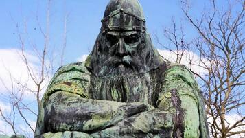 Una estatua de una figura con casco y barba, brazos cruzados, frente a un cielo despejado y árboles sin hojas.