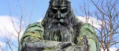 A statue of a figure with a helmet and beard, arms crossed, set against a clear sky and leafless trees.