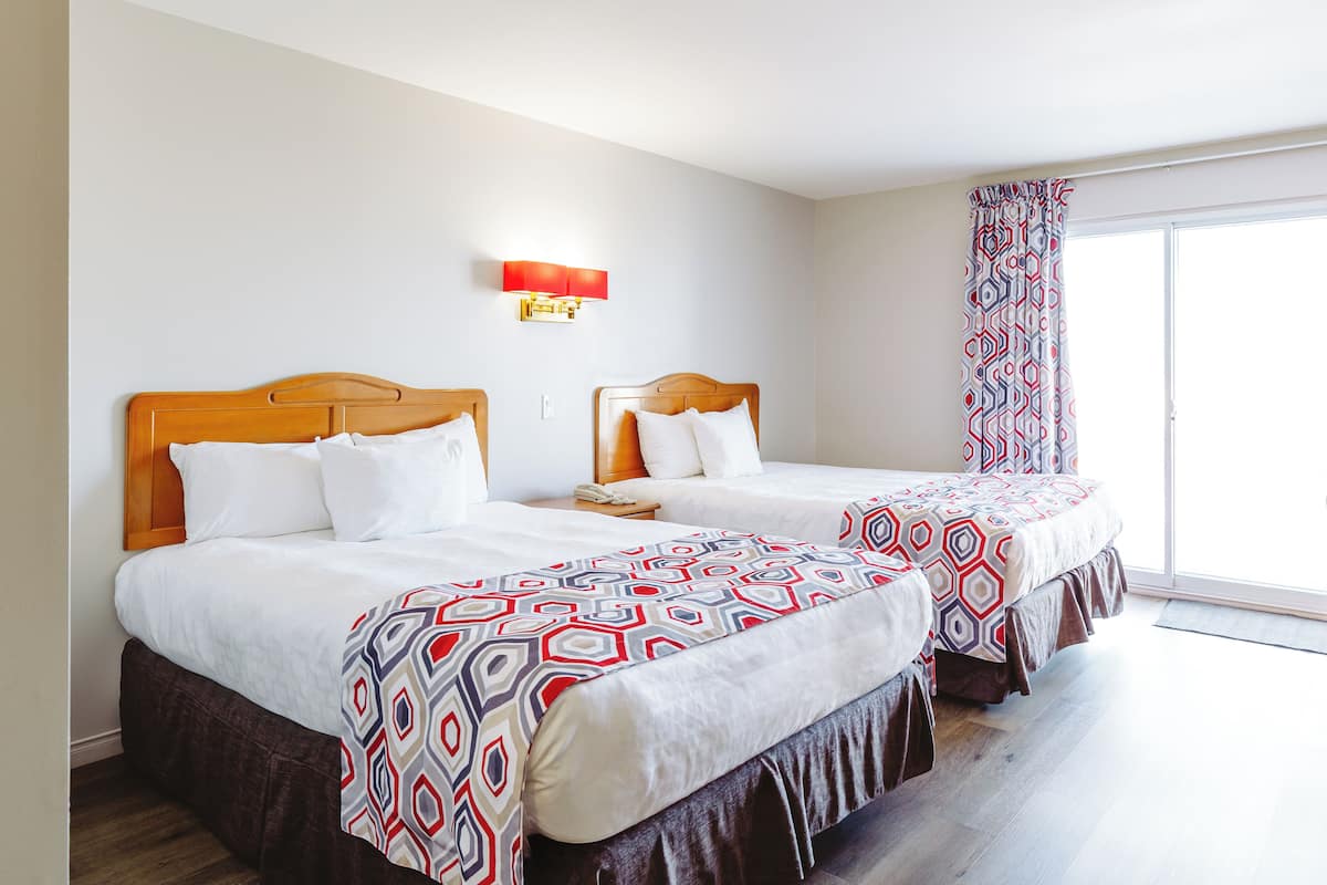 A hotel room with two beds, a nightstand with a phone, a wall-mounted light, and a large window with patterned curtains.