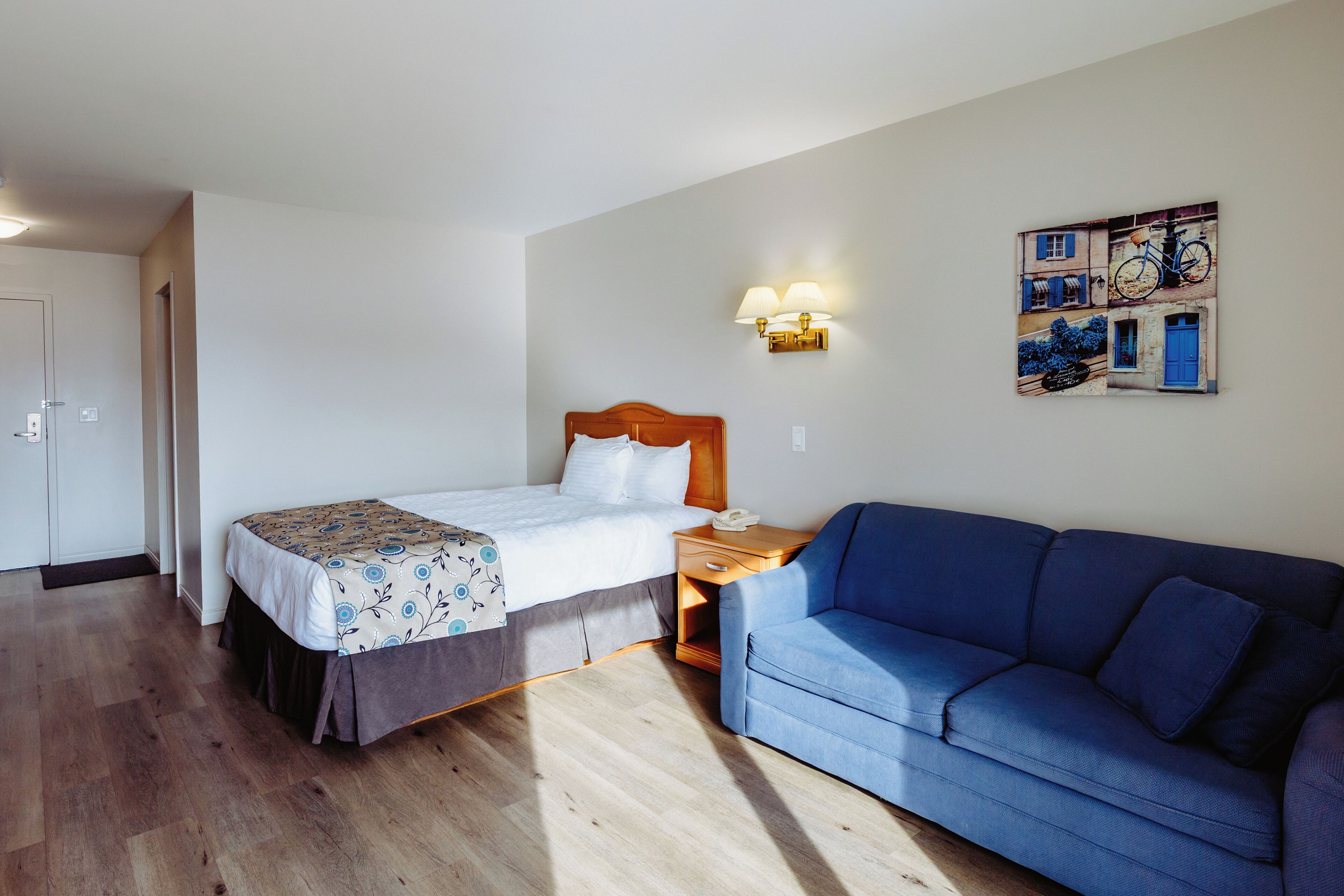 A hotel room with a bed, a blue sofa, a nightstand, a wall-mounted lamp, and a wall art depicting bicycles.