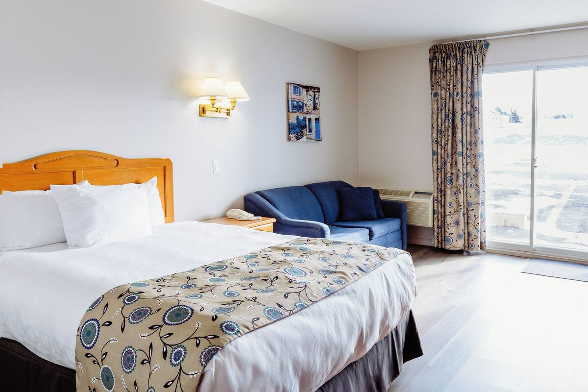 A hotel room with a bed, a desk, a chair, a large window with curtains, and a view of the outside.