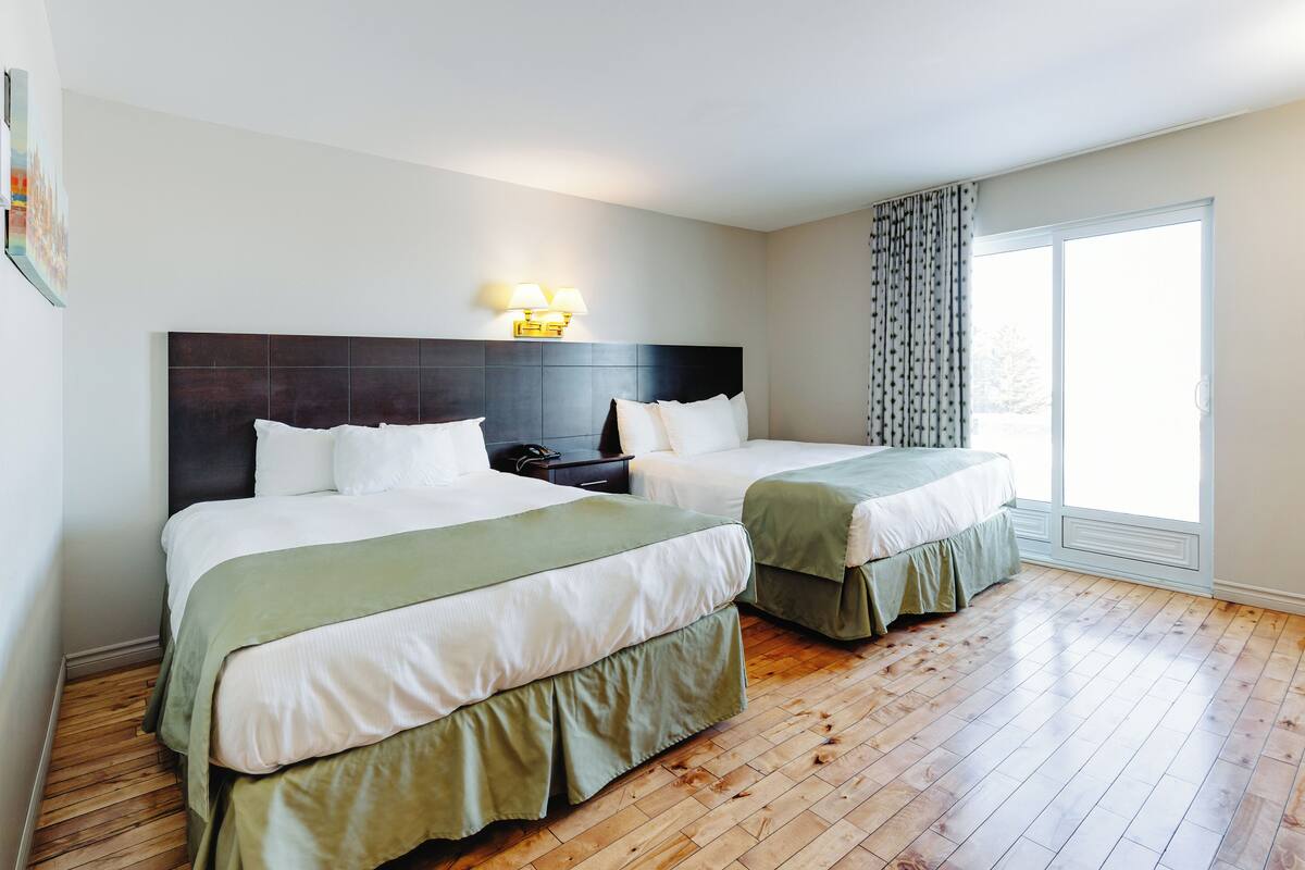 A hotel room with two beds, a dark headboard, bedside lamps, a nightstand, and a large window with curtains.