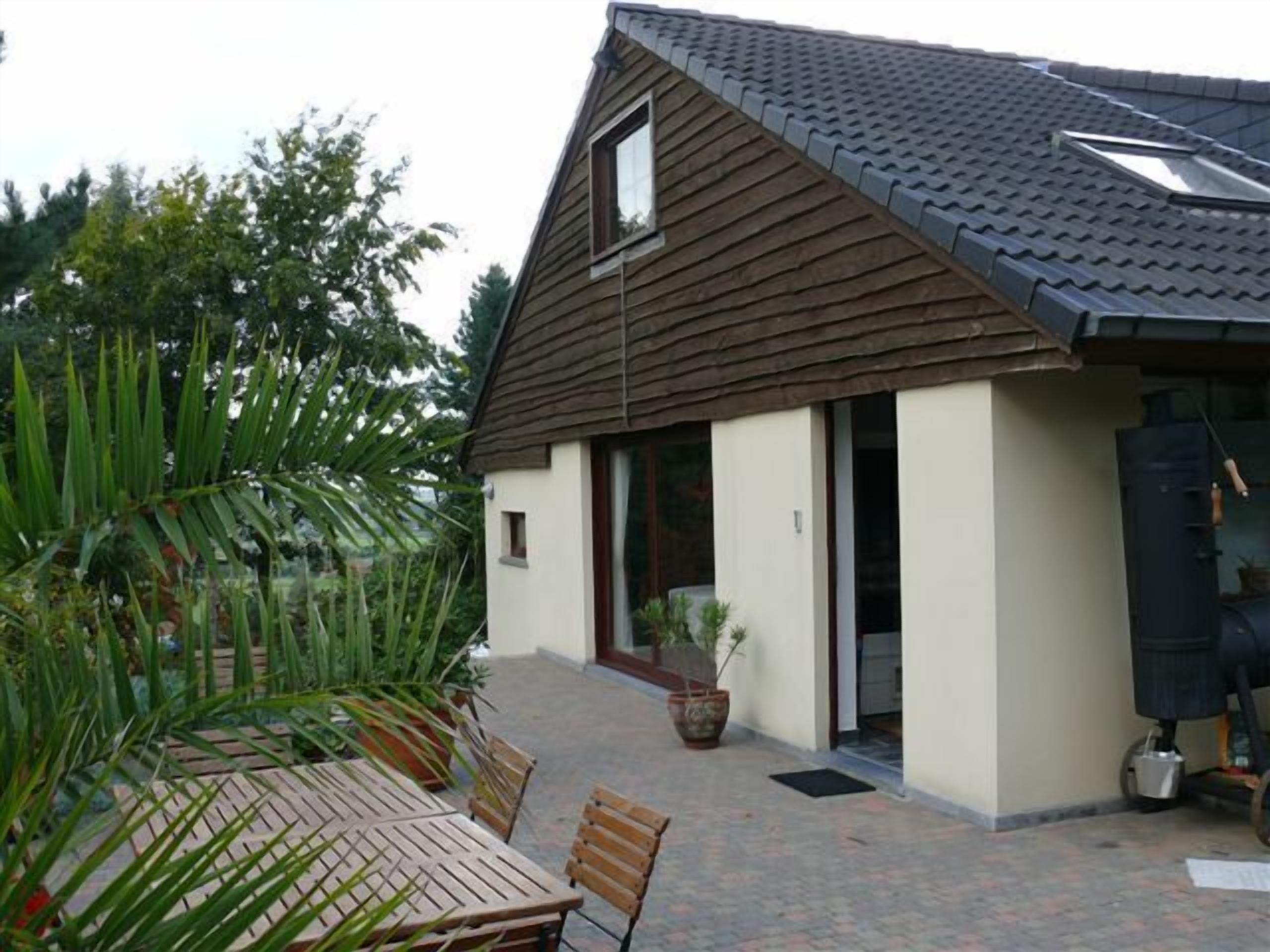 A house with a sloped roof, a small window, and a door leading to a covered patio area with a table and chairs.