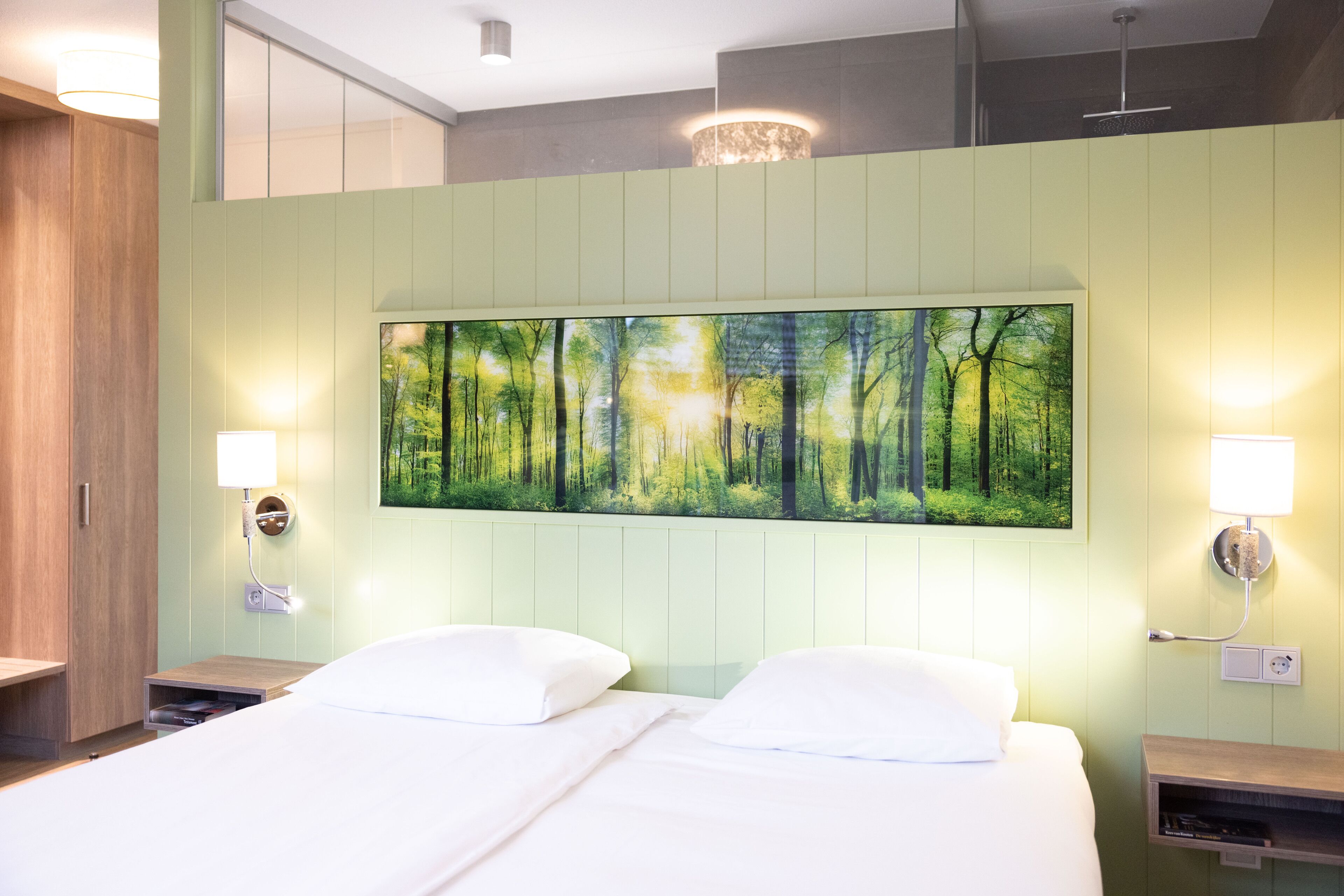 A hotel room with a bed, two bedside tables, a wall-mounted painting of a forest, and a ceiling light fixture.