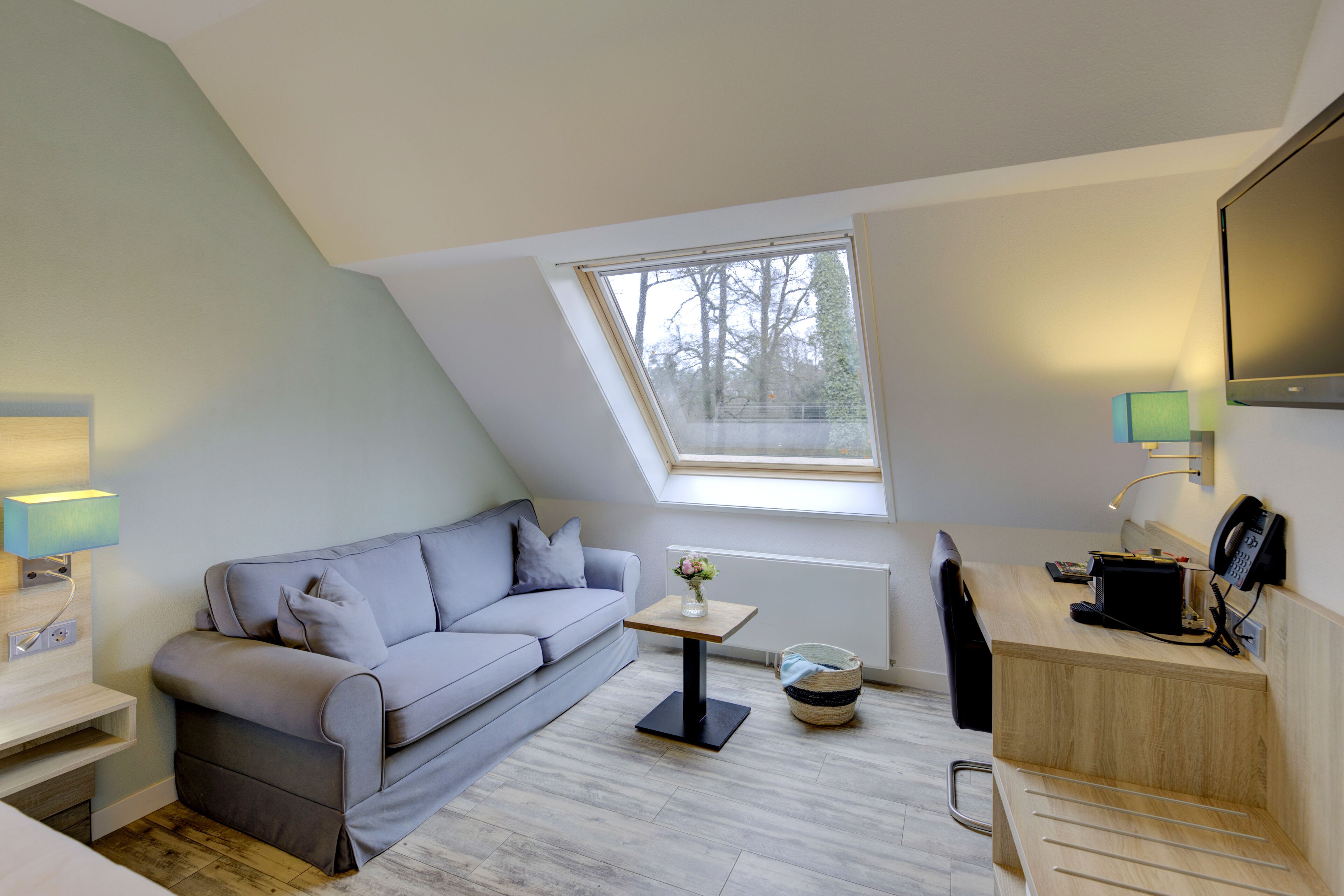 A modern, compact room with a sloped ceiling, a grey sofa, a small table with a vase, a desk with a lamp, and a large window providing natural light.