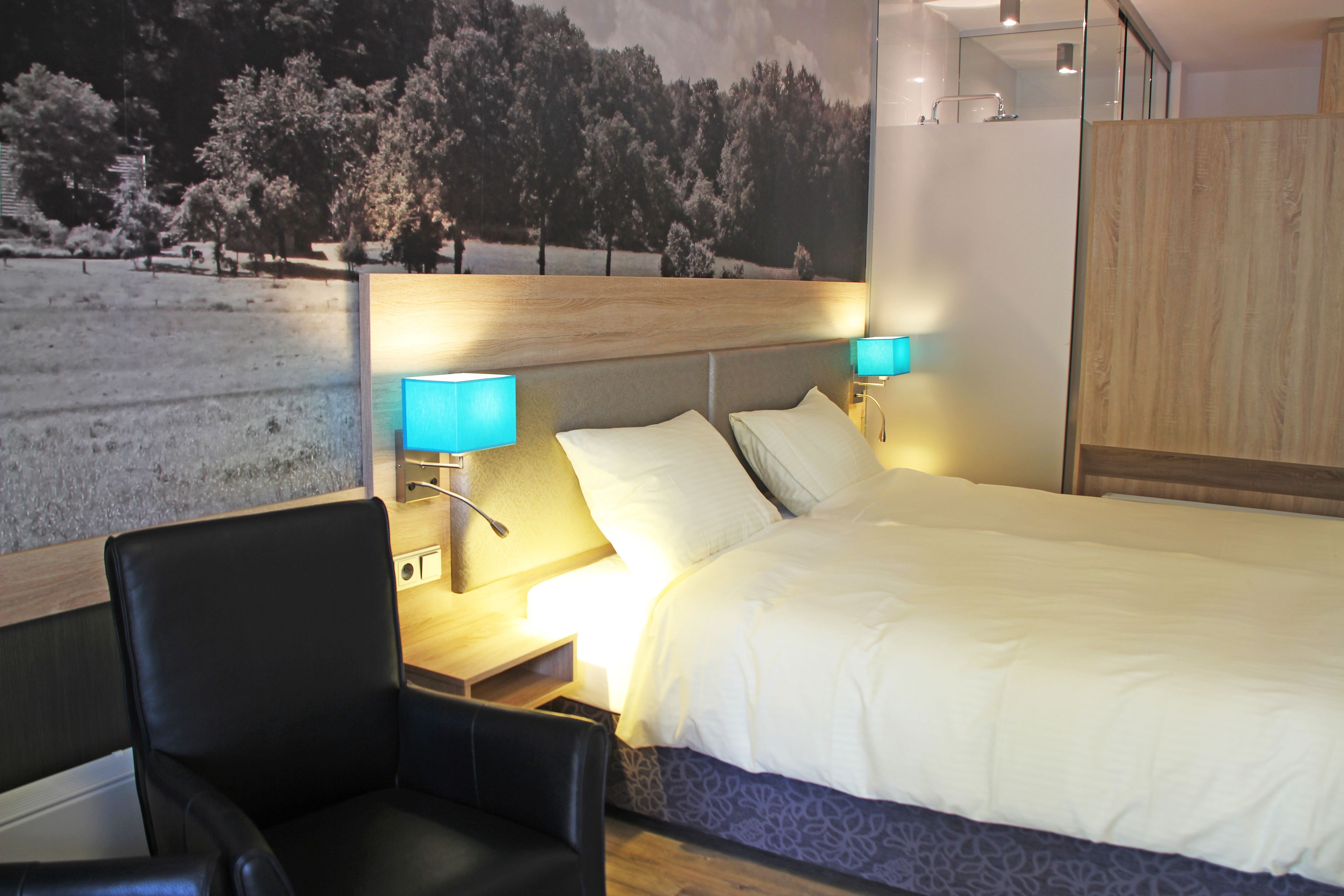 A modern hotel room with a large bed, two bedside lamps, a wooden headboard, a black leather chair, and a scenic wall mural.