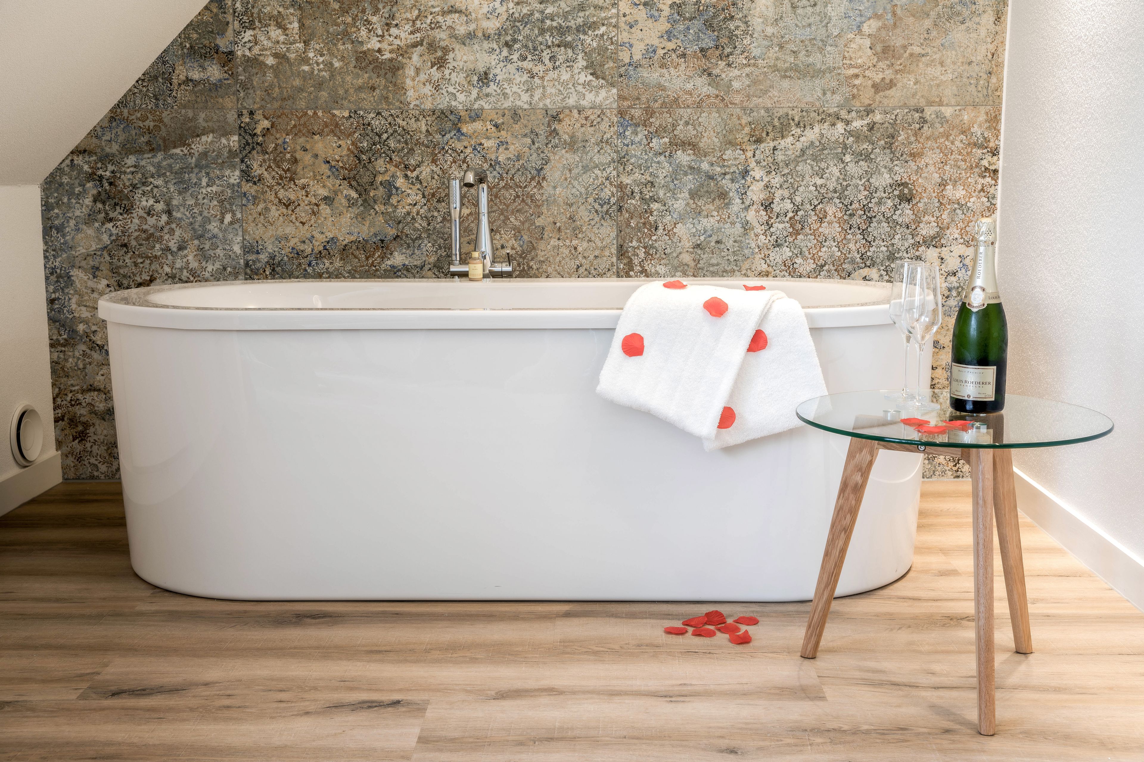 Une salle de bain moderne avec une baignoire sur pieds, une table en verre avec une bouteille et un verre, et une serviette blanche à pois rouges.