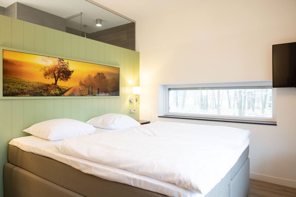 A bedroom with a bed, a painting of a sunset, a window, and a TV mounted on the wall.
