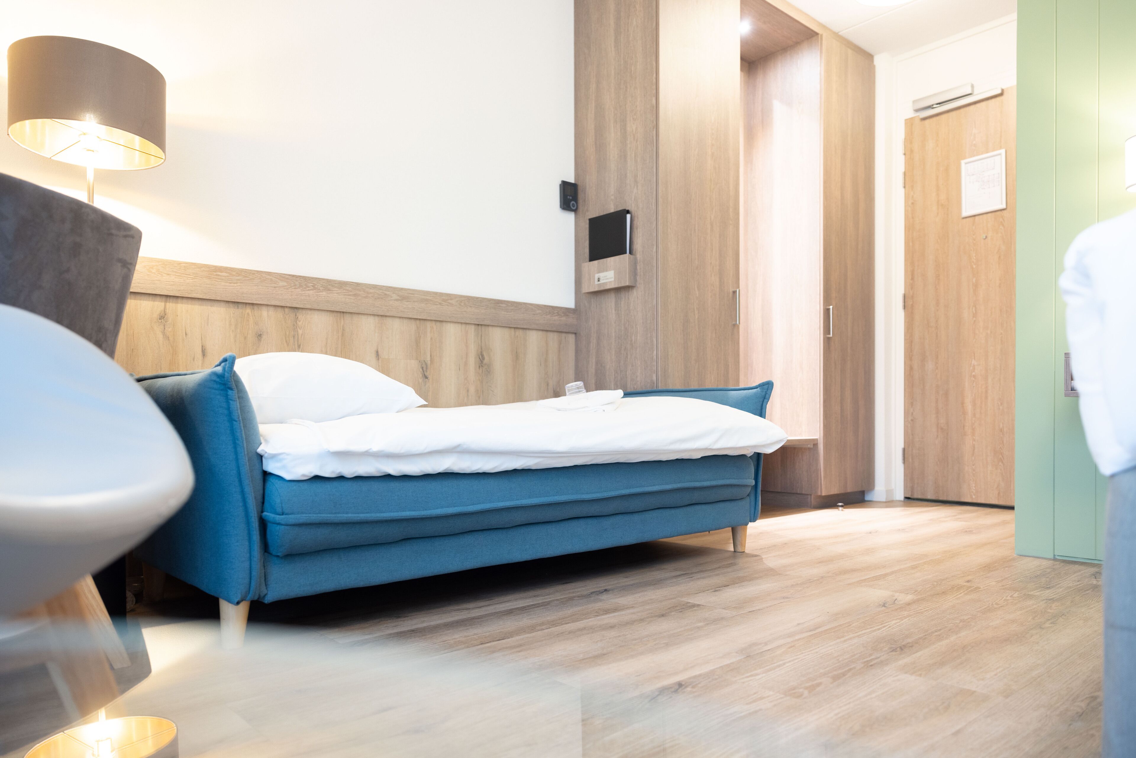 Una habitación de hotel moderna con una cama azul, suelo de madera, un televisor montado en la pared y una lámpara.