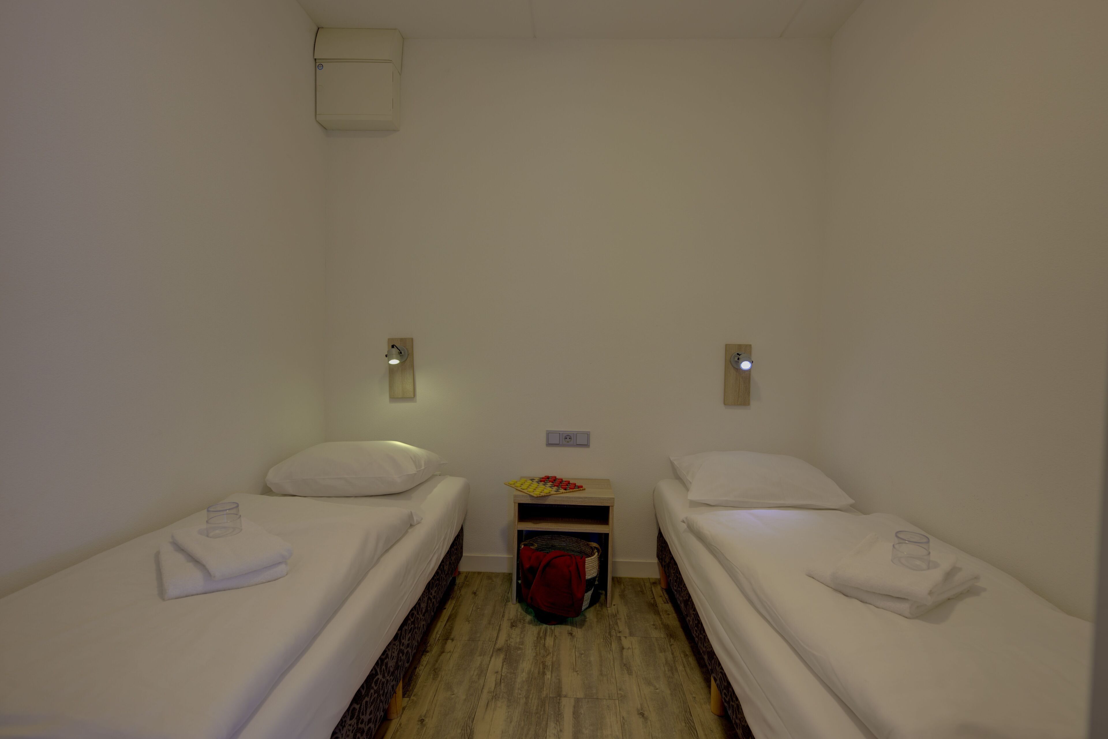 A small, clean room with two single beds, a nightstand, and a wall-mounted light fixture.