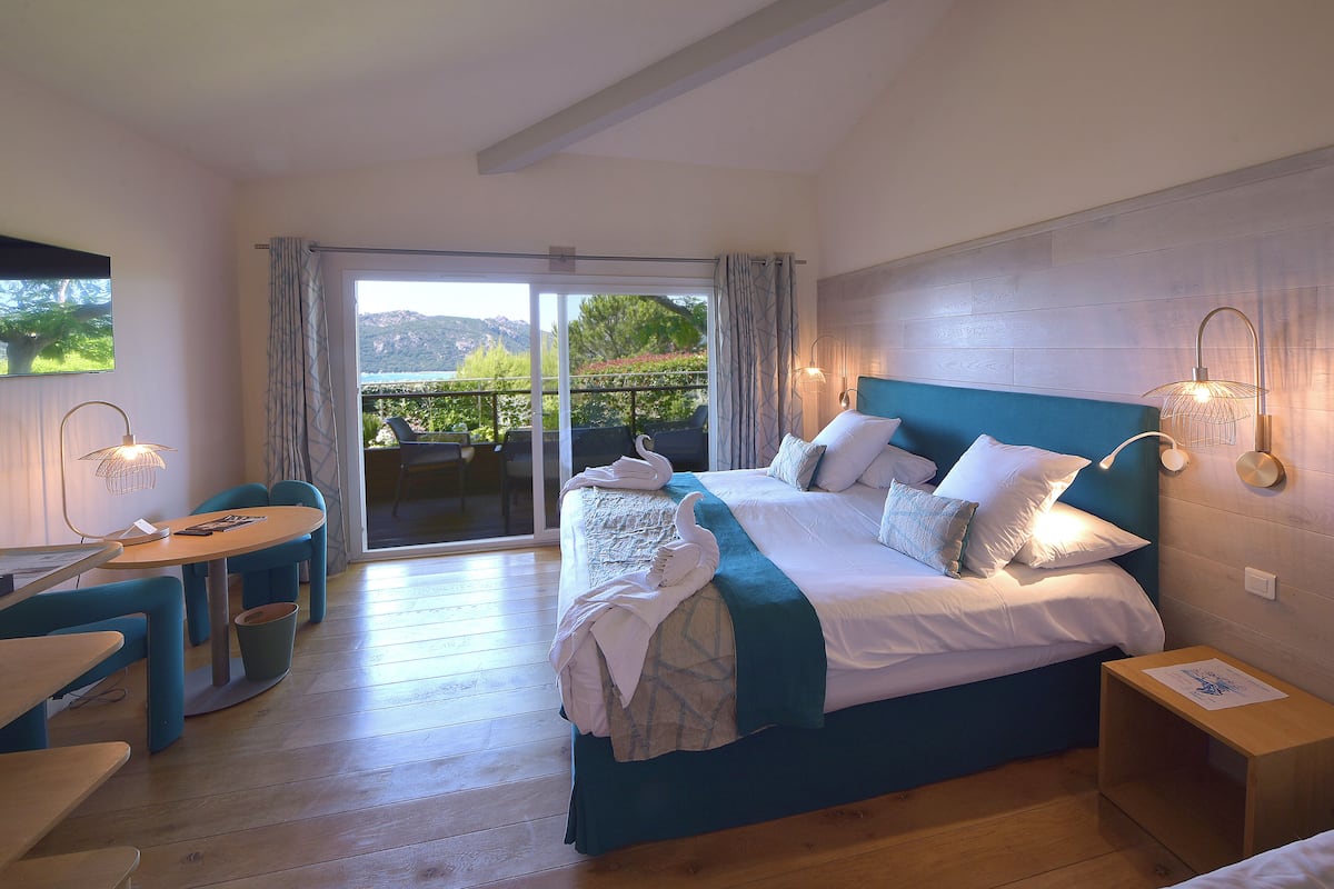 Un dormitorio con cama, escritorio y silla. Tiene un balcón con vistas a árboles y colinas.