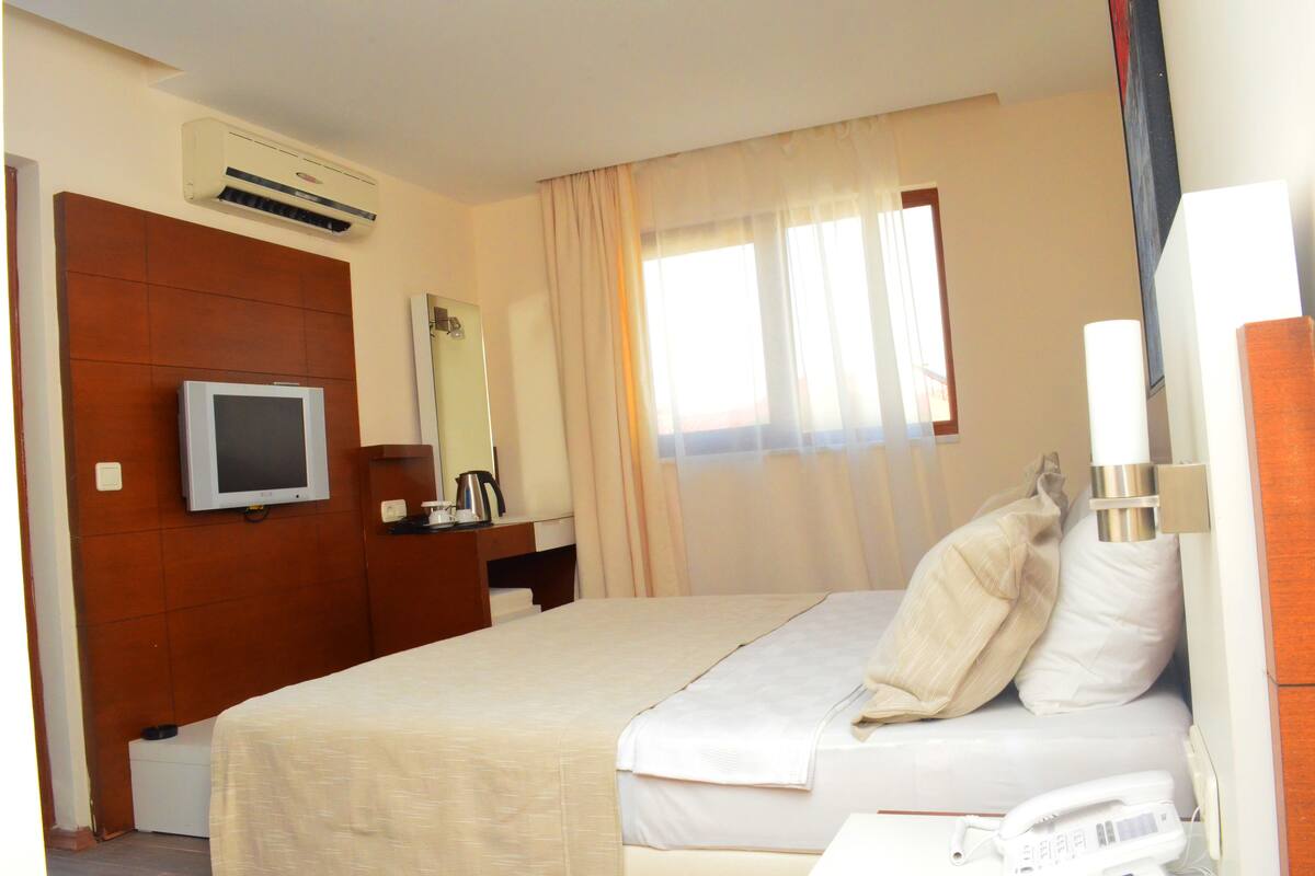 Una habitación de hotel con cama, televisión, espejo, ventana con cortinas y teléfono.