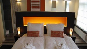 Una cama doble con sábanas blancas y dos almohadas, una mesita de noche con lámpara, y un aplique en la pared.
