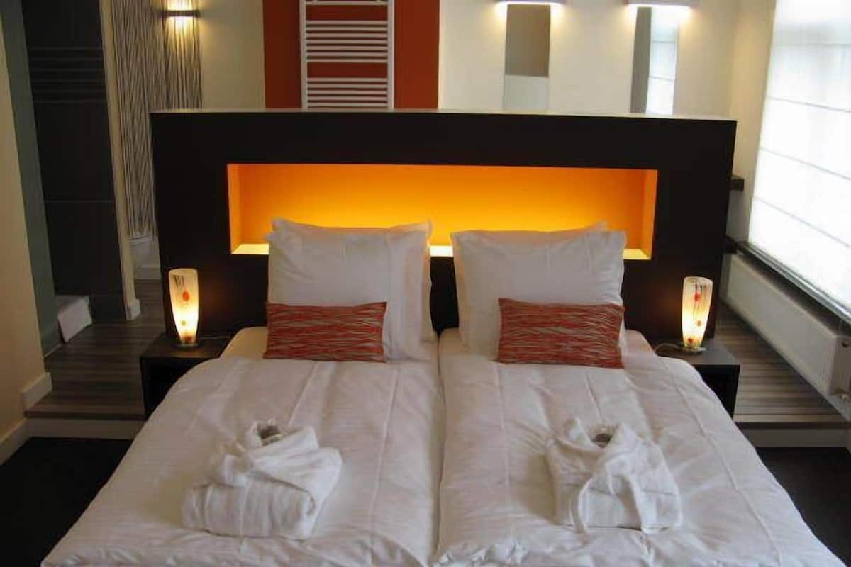 Una cama doble con sábanas blancas y dos almohadas, una mesita de noche con lámpara, y un aplique en la pared.