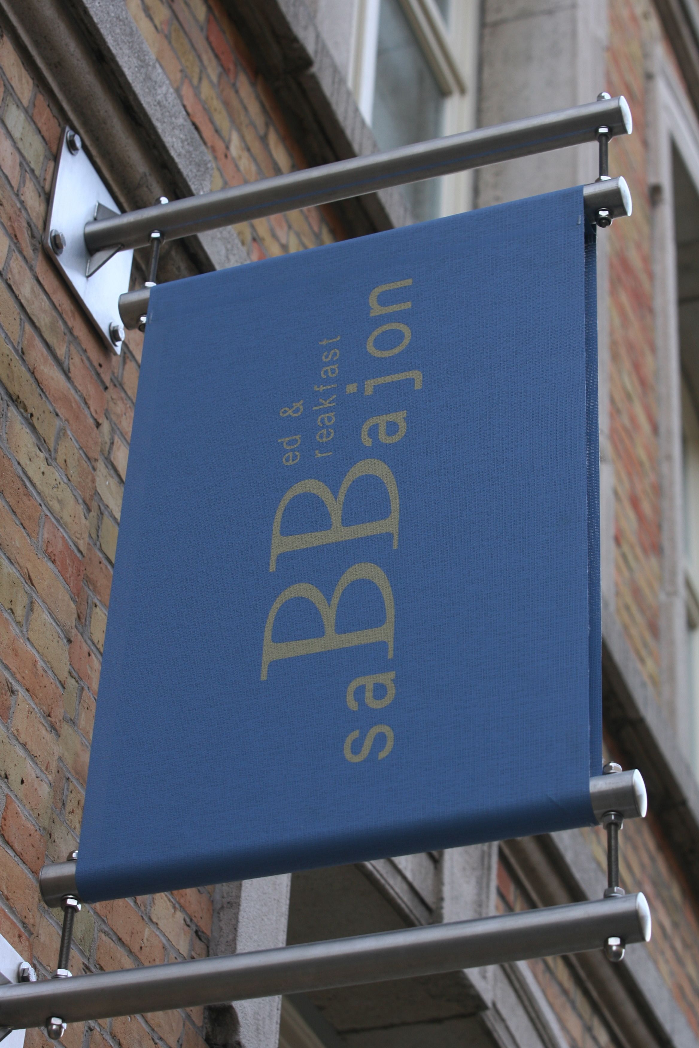 Ein blauer Banner mit dem Text "ed & breakfast" und "BB Bajon" an einer Metallhalterung vor einer Ziegelwand.