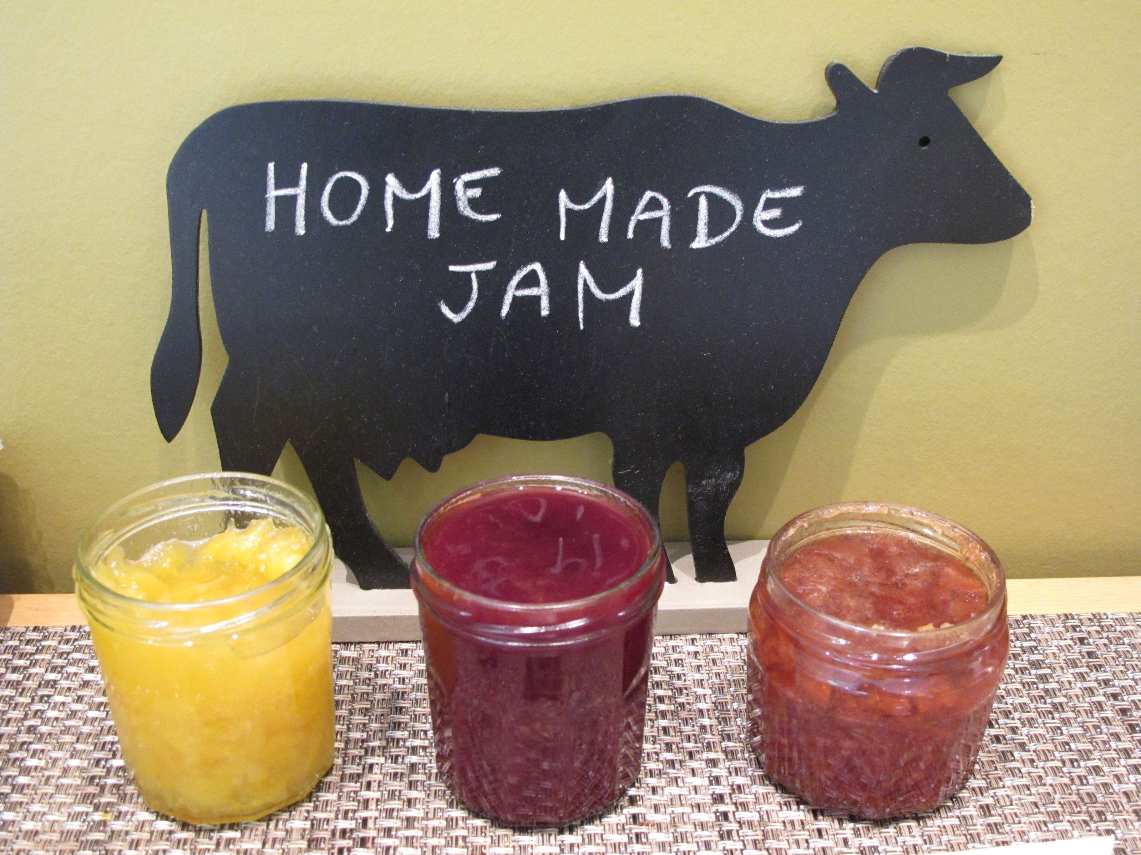 Three jars of jam on a woven mat with a chalkboard sign reading "HOME MADE JAM" behind them.