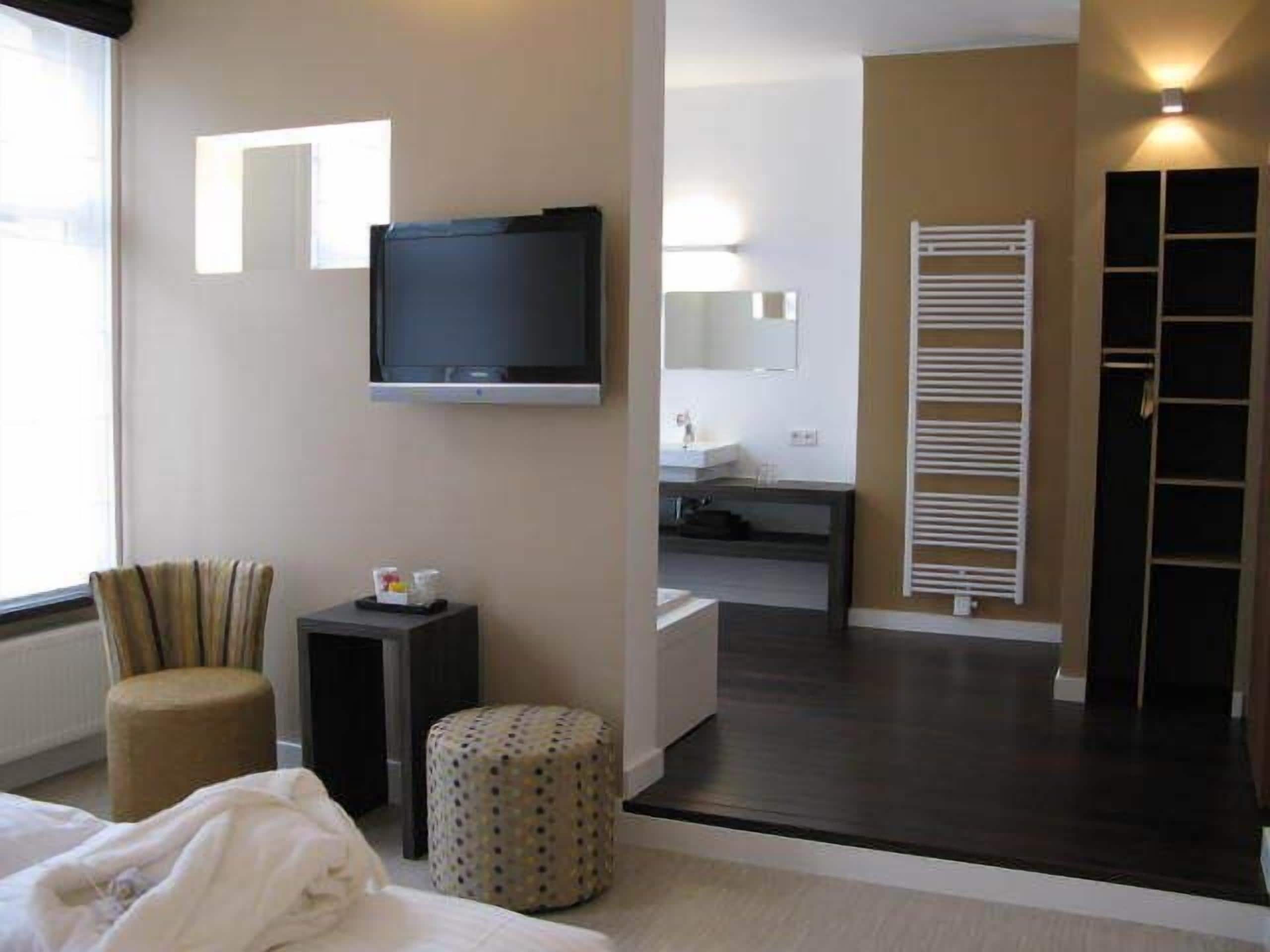 A hotel room with a bed, a TV, a chair, a small table, a round ottoman, a bathroom with a sink and mirror, and a wardrobe.