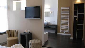 A hotel room with a bed, a TV, a chair, a small table, a round ottoman, a bathroom with a sink and mirror, and a wardrobe.