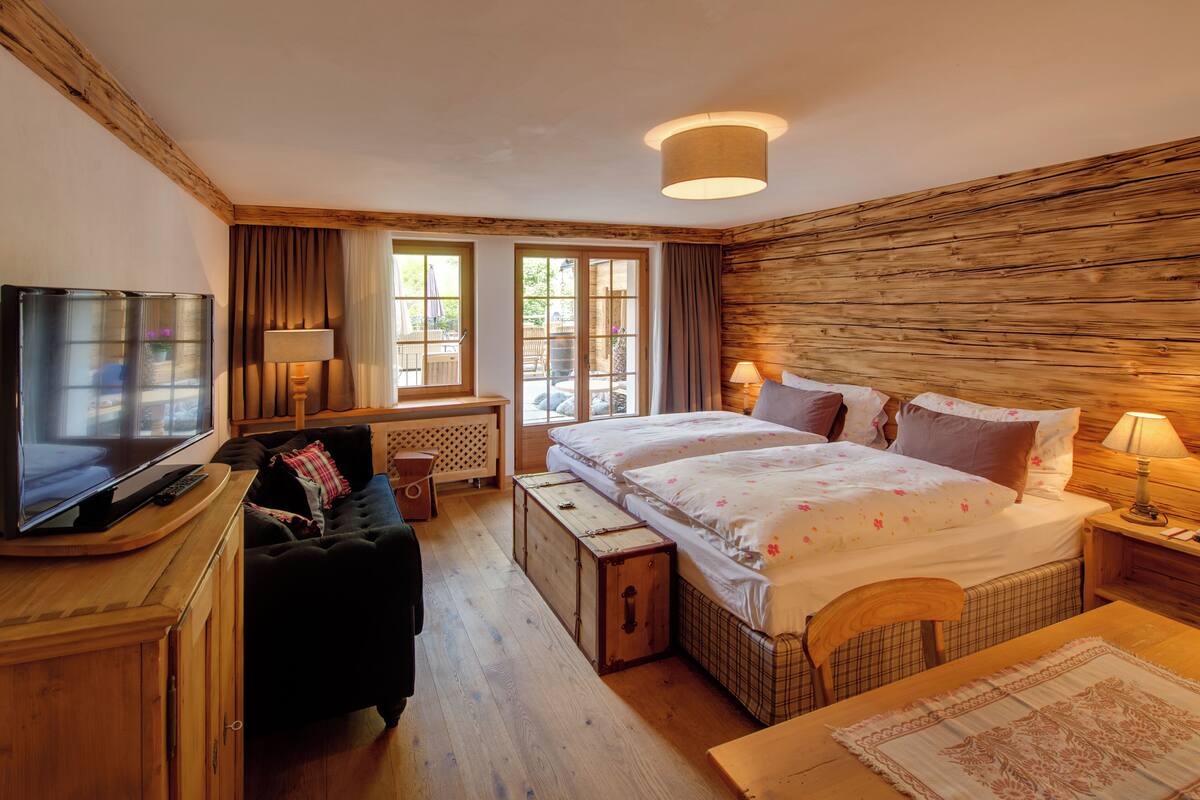 Quarto aconchegante com teto de madeira, uma cama grande com roupa de cama floral, uma televisão em um armário de madeira e uma janela com cortinas.
