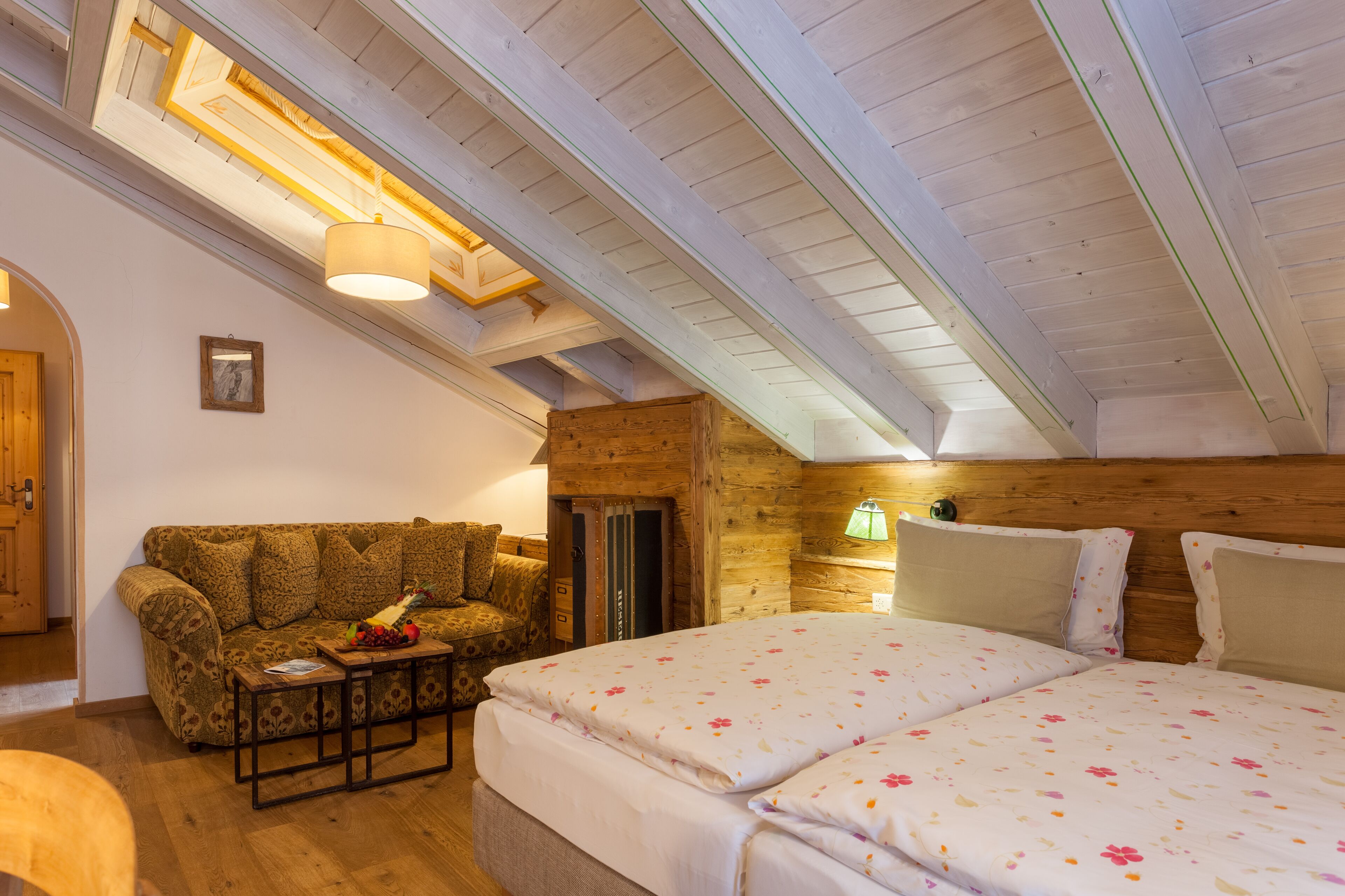 A cozy room with a wooden ceiling, a bed with floral bedding, a sofa with a fruit basket, and a small table.
