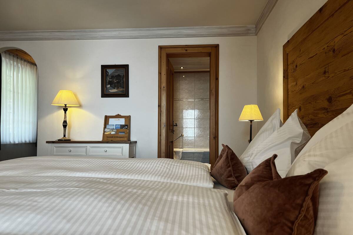 Quarto de hotel com uma cama grande, duas luminárias de cabeceira, um painel de cabeceira de madeira, uma mesa de cabeceira com uma luminária, um letreiro de boas-vindas emoldurado e uma porta com painel de vidro.