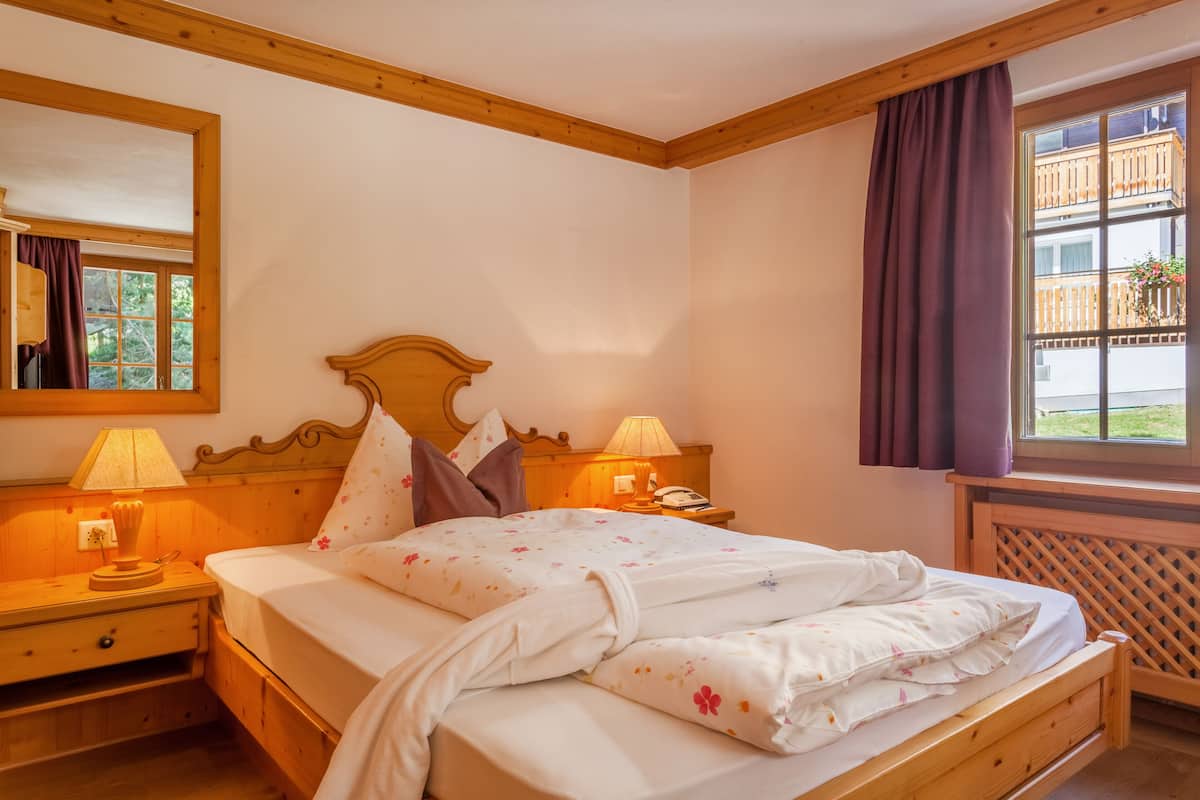 Cama bem arrumada com roupa de cama floral, cabeceira de madeira, abajures e uma janela com vista para uma varanda.