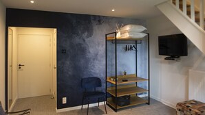 Una sala de estar moderna con una escalera, una pared de acento oscura, un área de descanso con una silla, un televisor montado en la pared y una mesita con una lámpara.