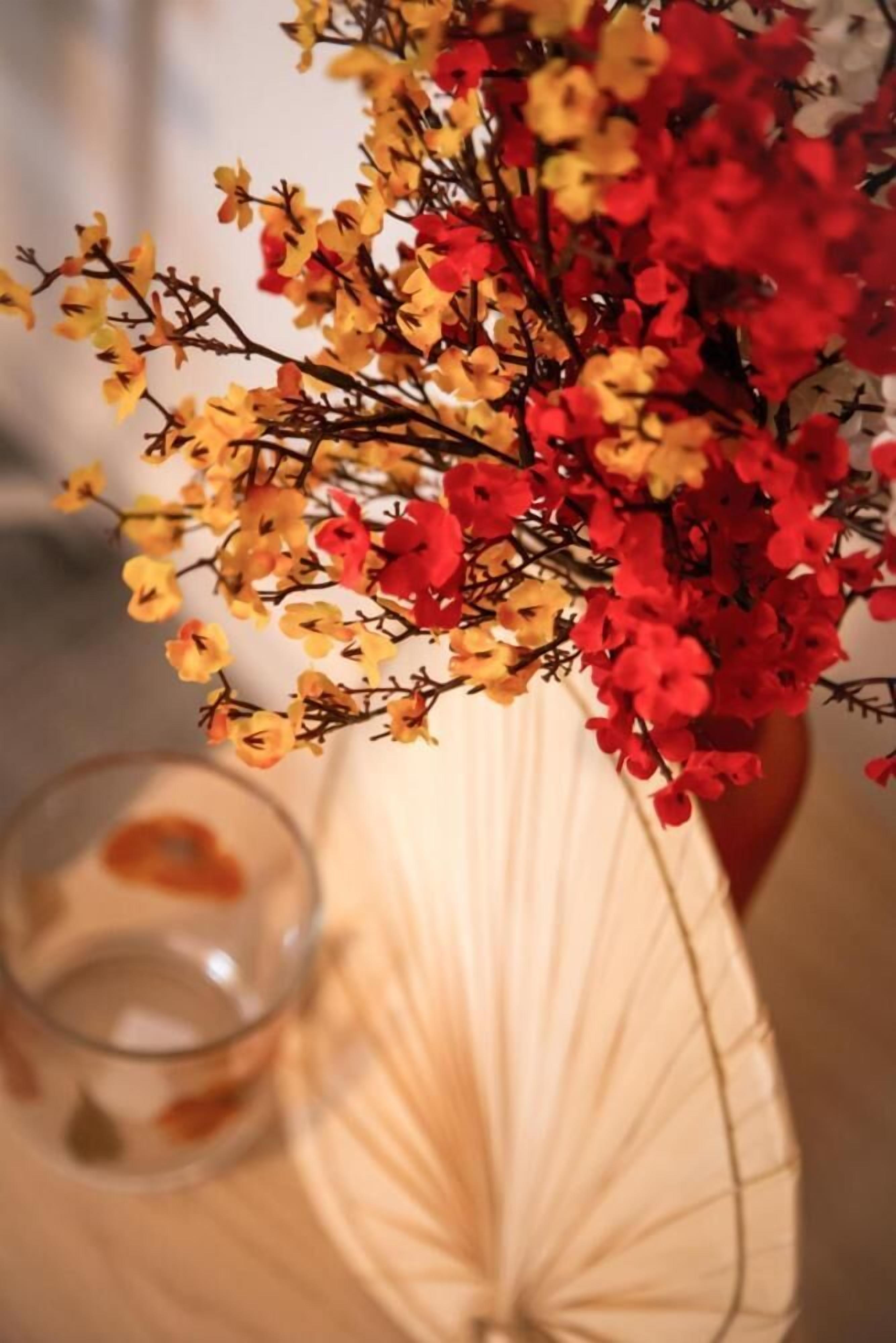 Um vaso com flores vermelhas e amarelas, um copo com bebida e um leque branco sobre uma mesa.