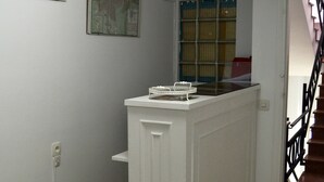 A hallway with a white baseboard, a small white shelf, a framed picture, and a window with blue and yellow curtains. - Hostal Isabel II (Figueres)
