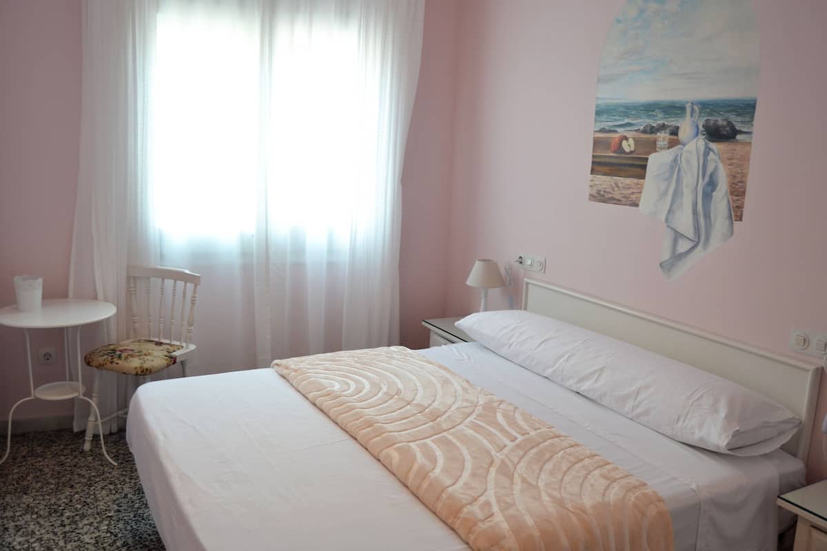 A bedroom with a bed, a chair, a small table, and a painting of a beach scene.