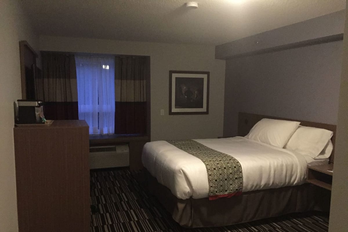A hotel room with a bed, a desk, a window with curtains, a framed picture on the wall, and a patterned carpet.