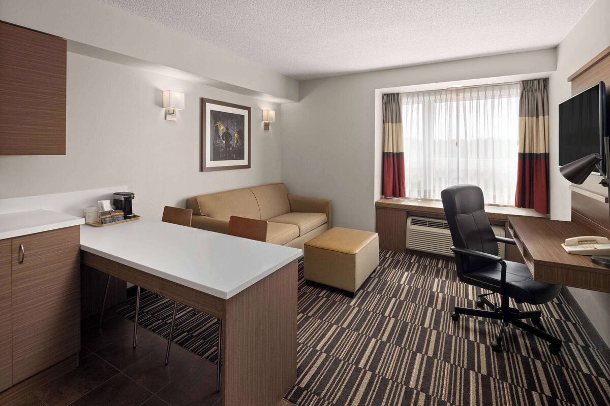A modern hotel room with a white desk, a beige sofa, a black office chair, a flat-screen TV, and a window with curtains.