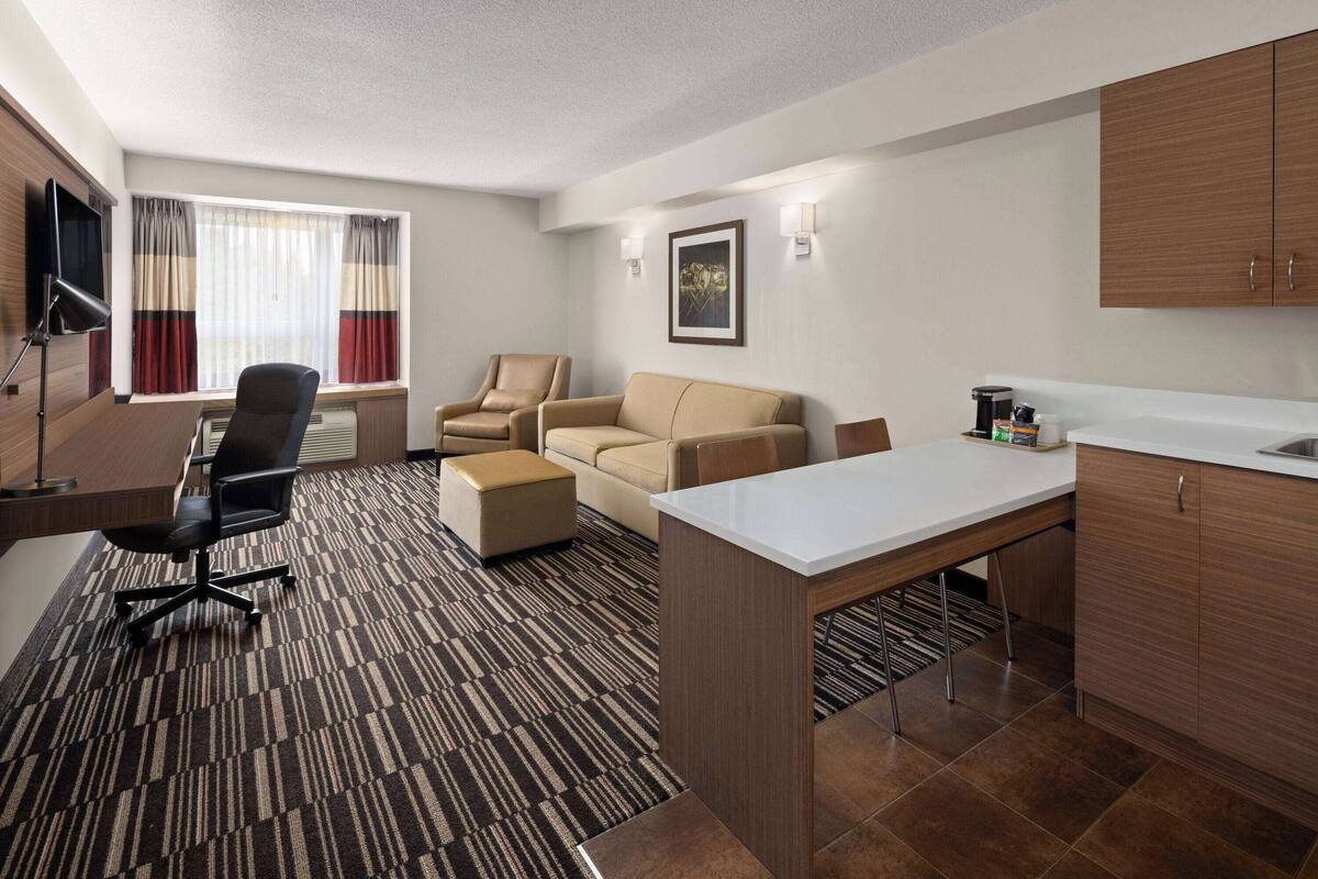 A modern hotel room with a desk, office chair, sofa, and kitchenette.
