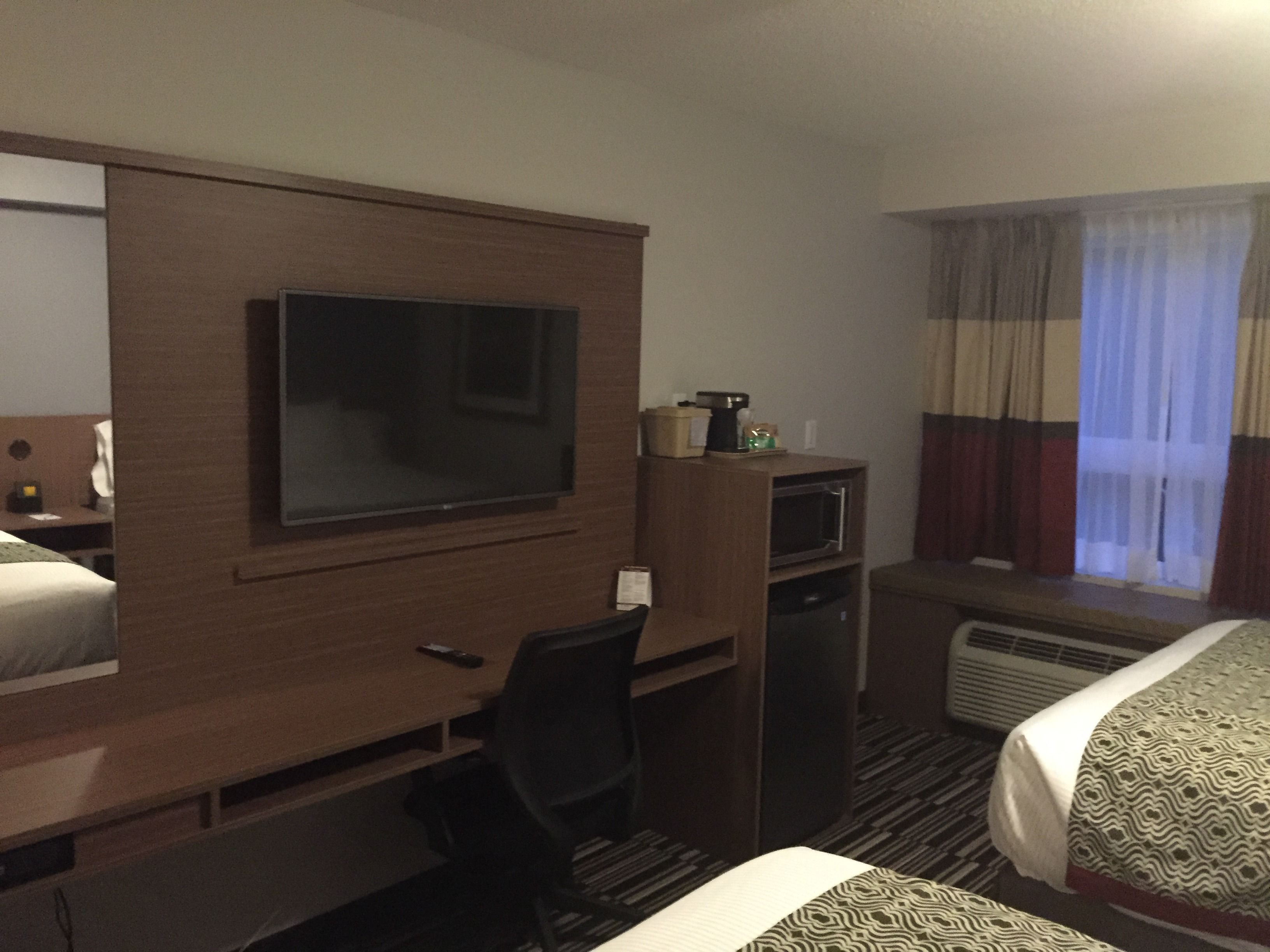 Une chambre d’hôtel comprenant un lit, un bureau avec une télévision, une chaise, un four à micro-ondes et une fenêtre avec des rideaux.