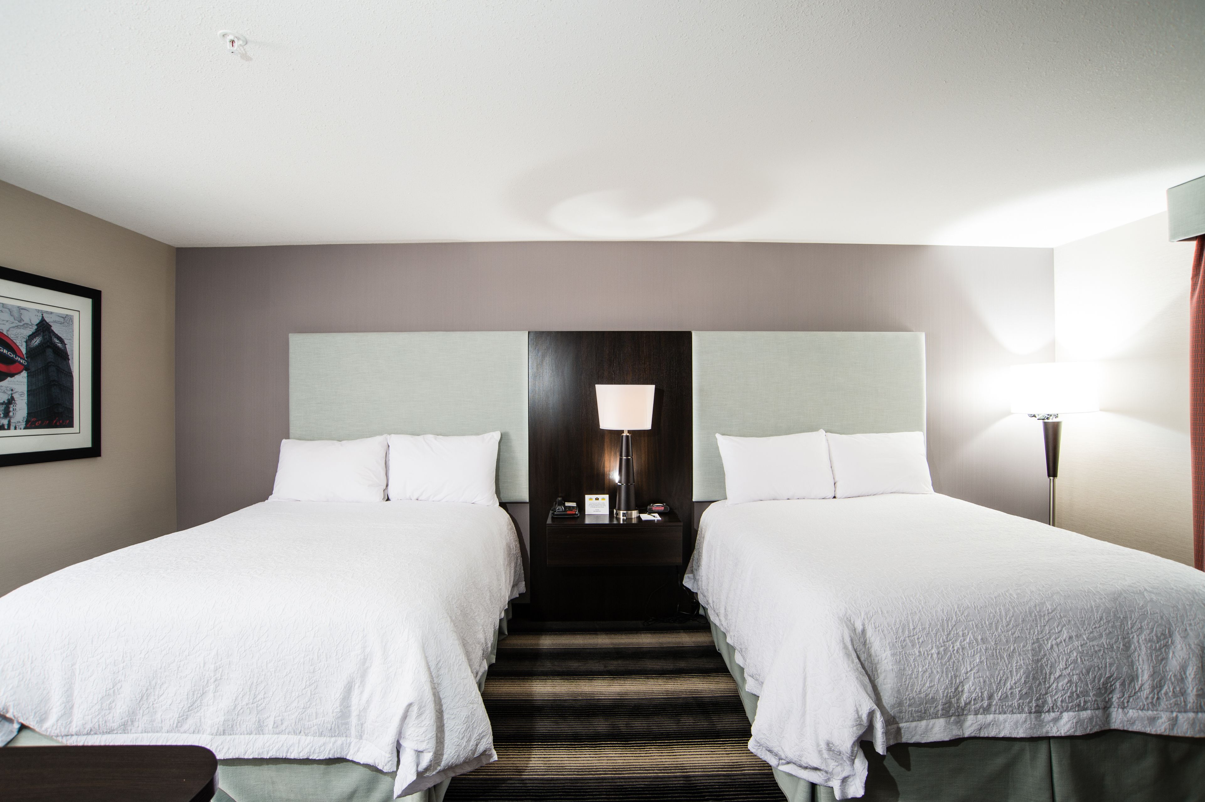 A hotel room with two beds, a nightstand with a lamp, and a framed picture on the wall.