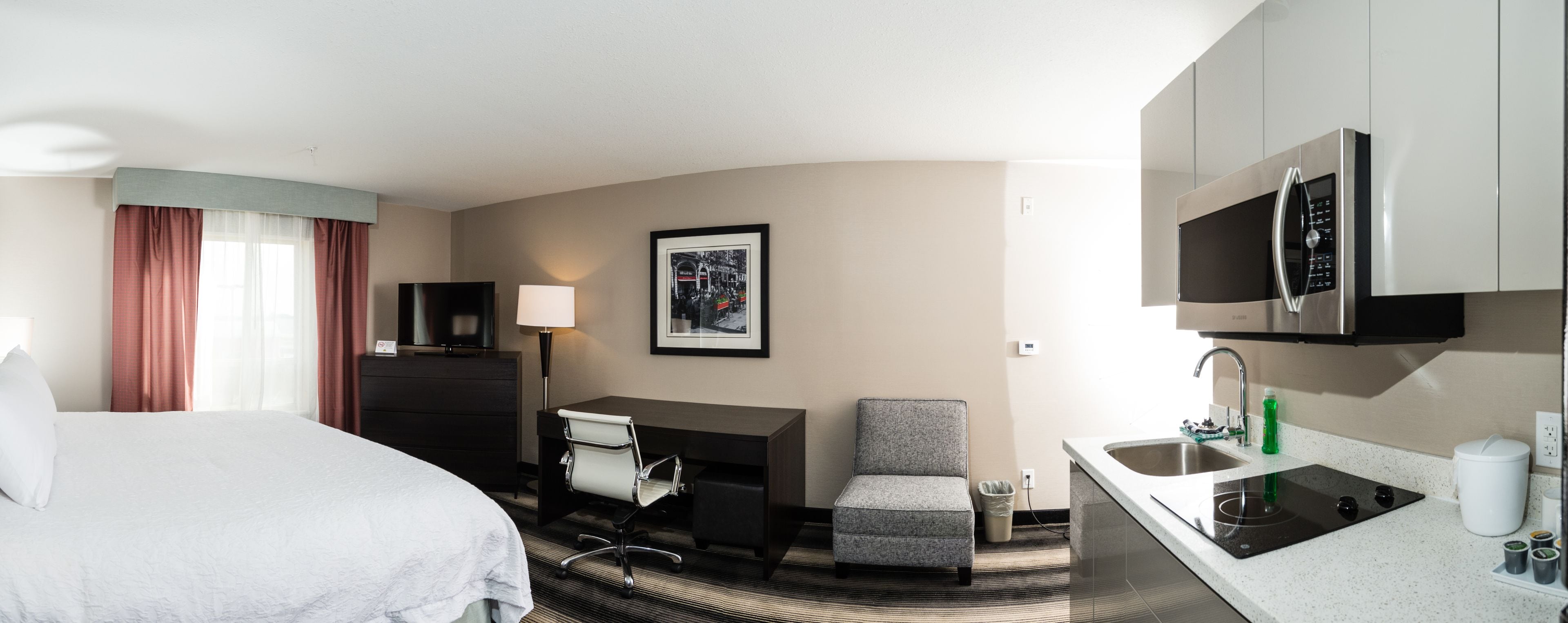 A hotel room with a bed, desk, chair, TV, and kitchenette.