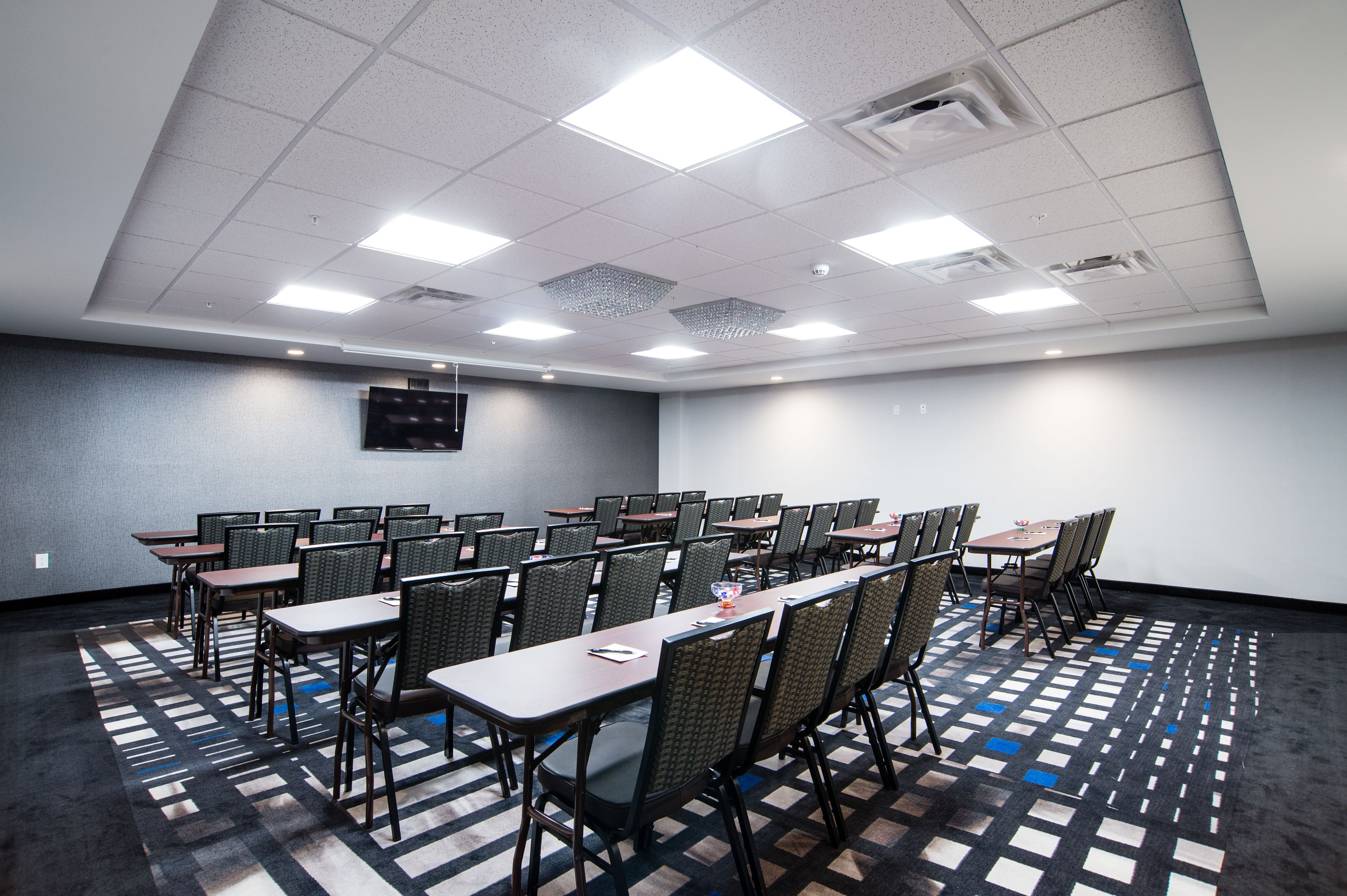 A conference room with multiple tables and chairs, a flat-screen TV mounted on the wall, and a patterned carpet.