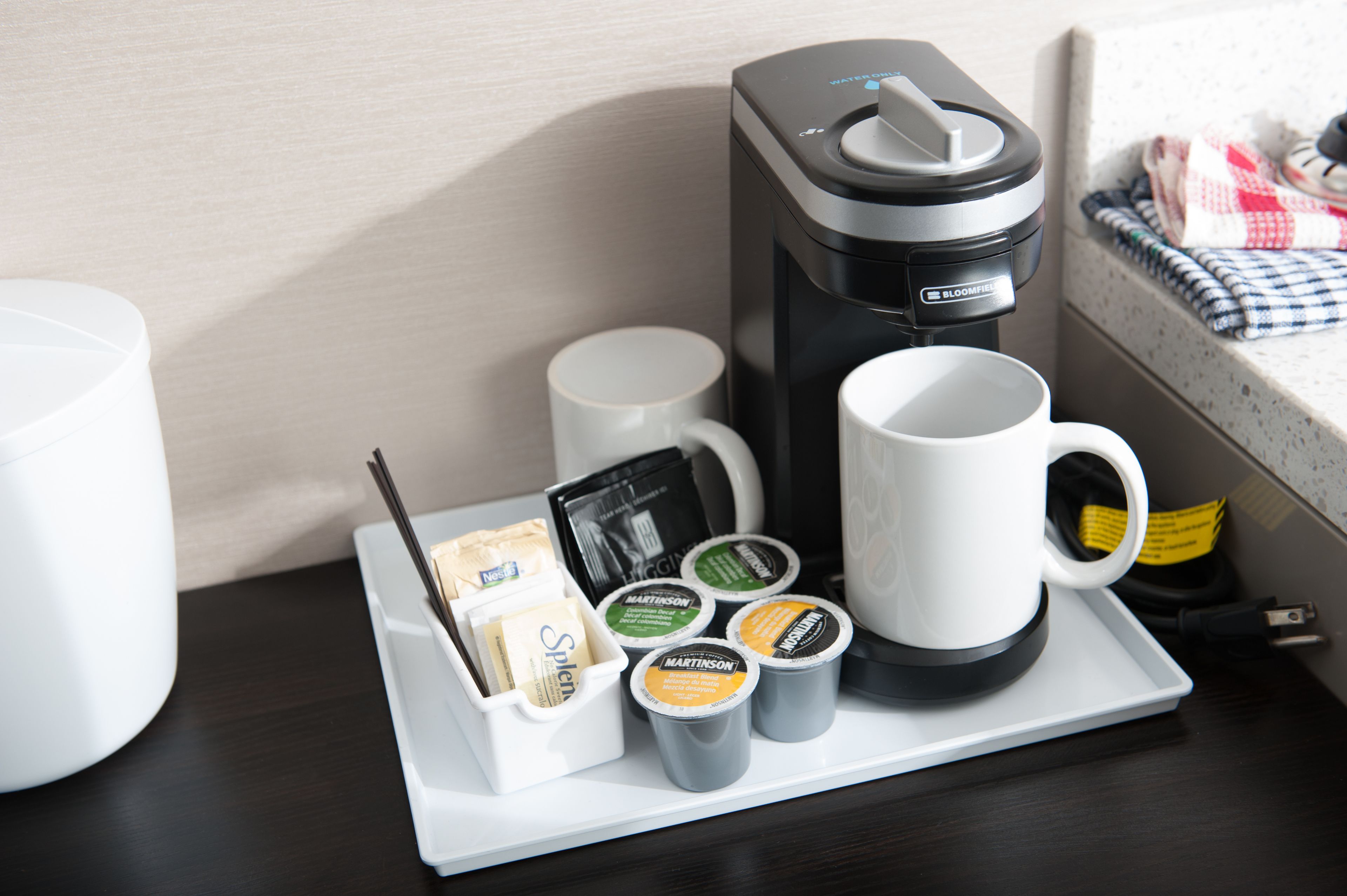 Une machine à café, une tasse blanche, une soucoupe blanche et un plateau contenant des dosettes de café et une cuillère, le tout posé sur une table en bois foncé.