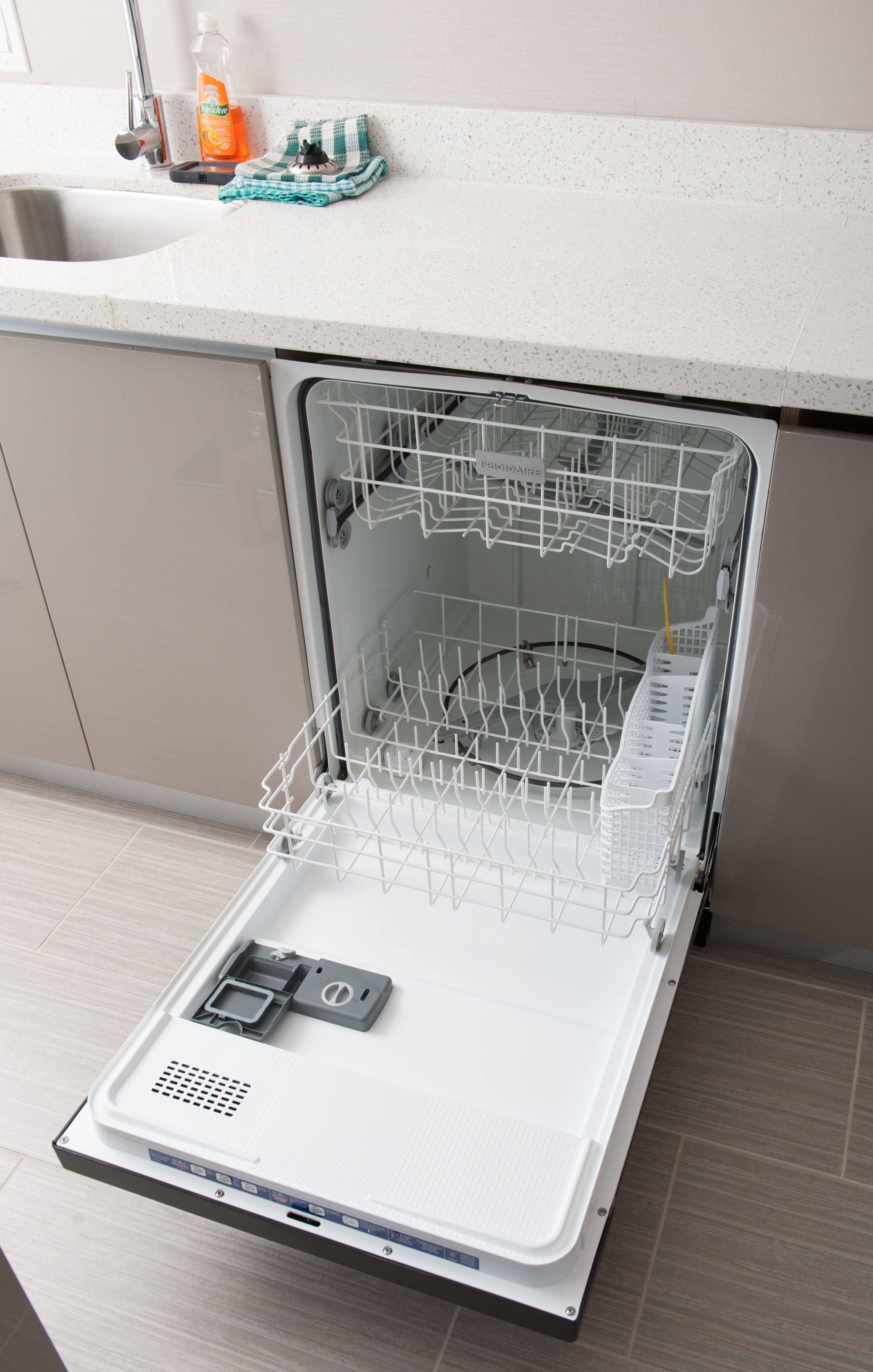 An open dishwasher with a detergent bottle and a cloth on the countertop above it.