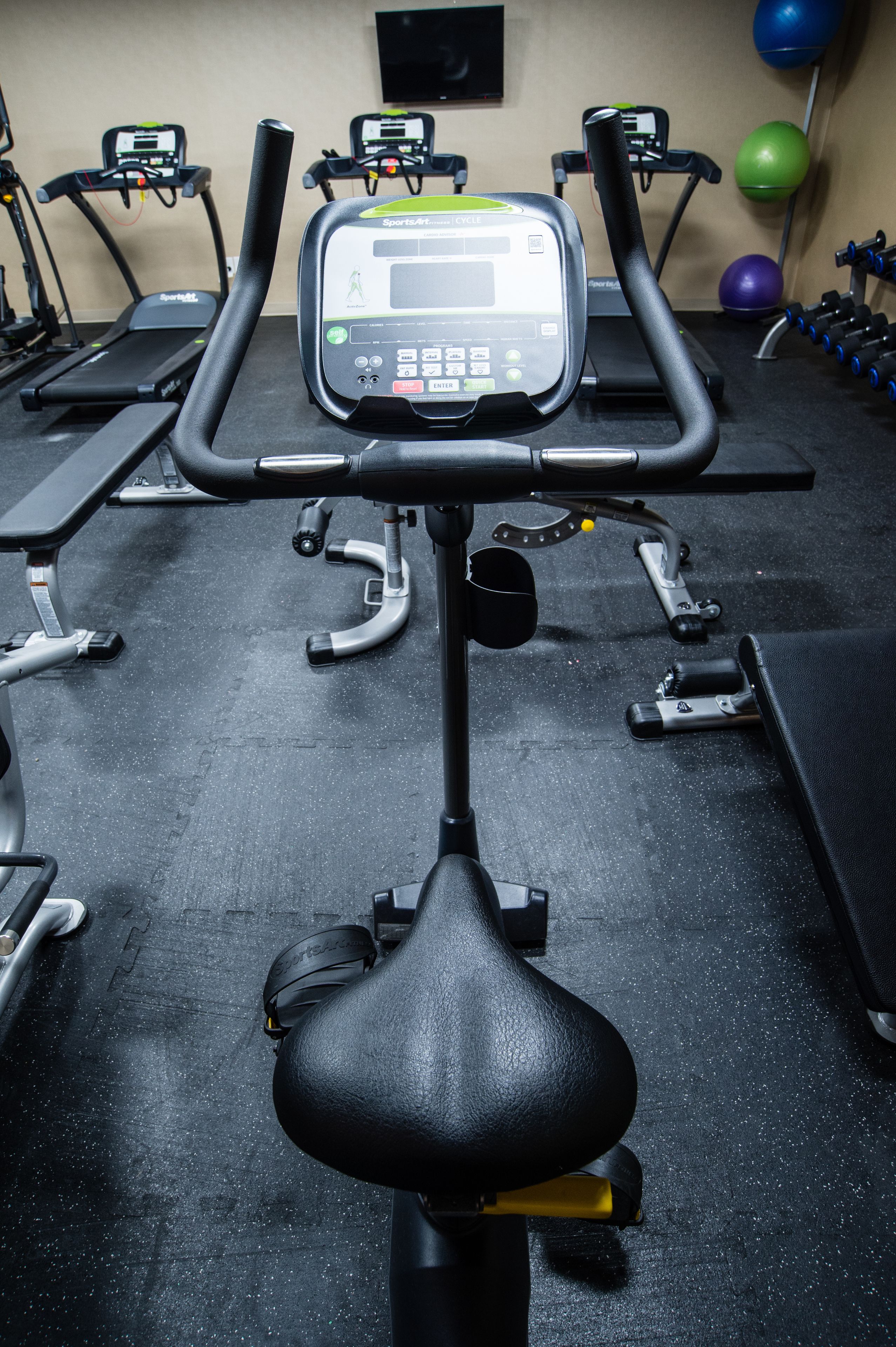 Una bicicleta estática con pantalla digital, un banco y pelotas de ejercicios en un gimnasio.