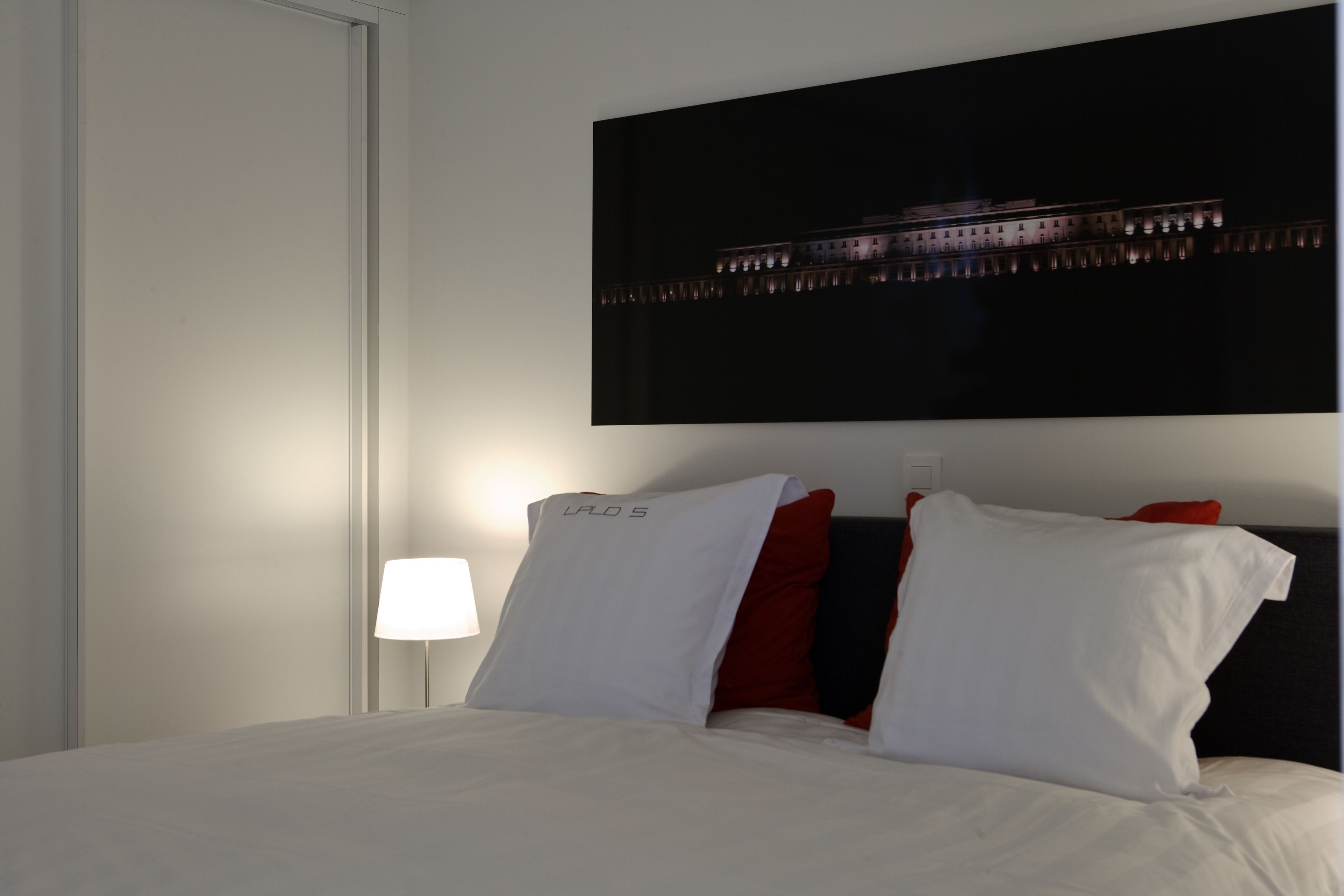 A hotel room with a bed, white pillows, a red pillow, a nightstand with a lamp, and a large framed picture on the wall.