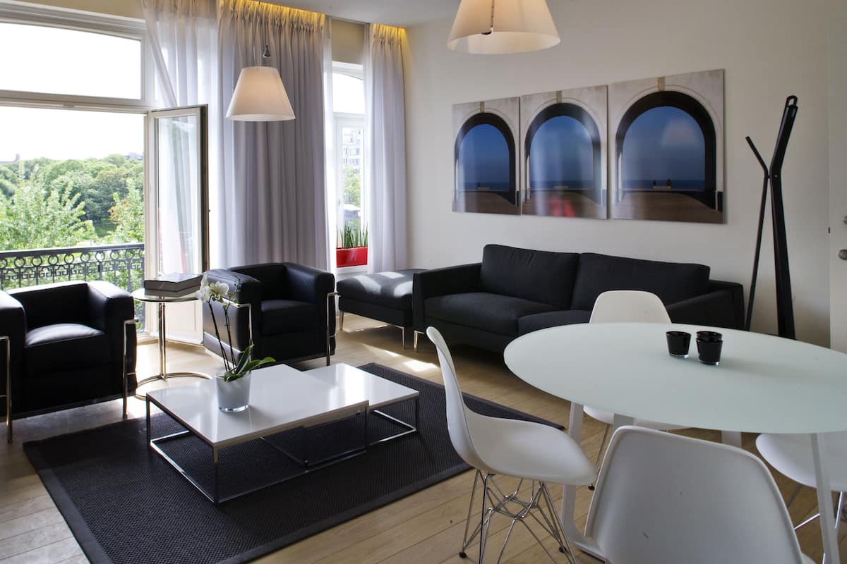 Un salón moderno con un sofá negro, sillas blancas, una mesa de centro y vistas de la ciudad a través de grandes ventanales.