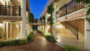 Pasillo de hotel de varios niveles con suelo de ladrillo, columnas blancas y balcones con barandillas de madera.