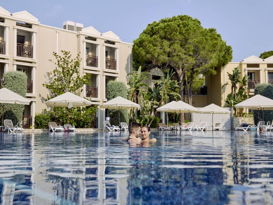 Una piscina con sillas de descanso y sombrillas, rodeada de edificios de varios pisos con balcones.