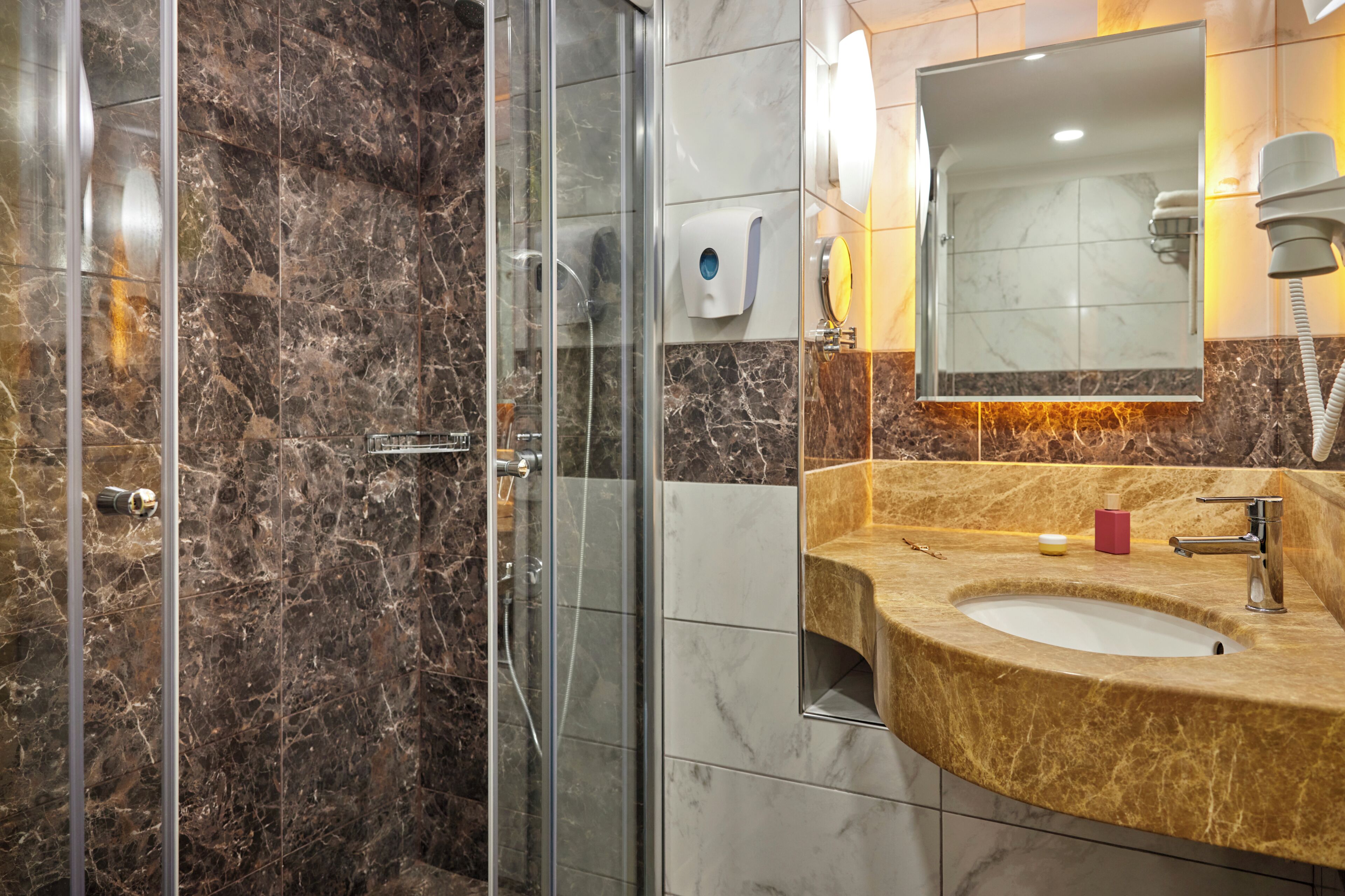 Un baño con ducha envuelta en vidrio, pared con azulejos de mármol, lavamanos con encimera de mármol y espejo sobre el lavamanos.